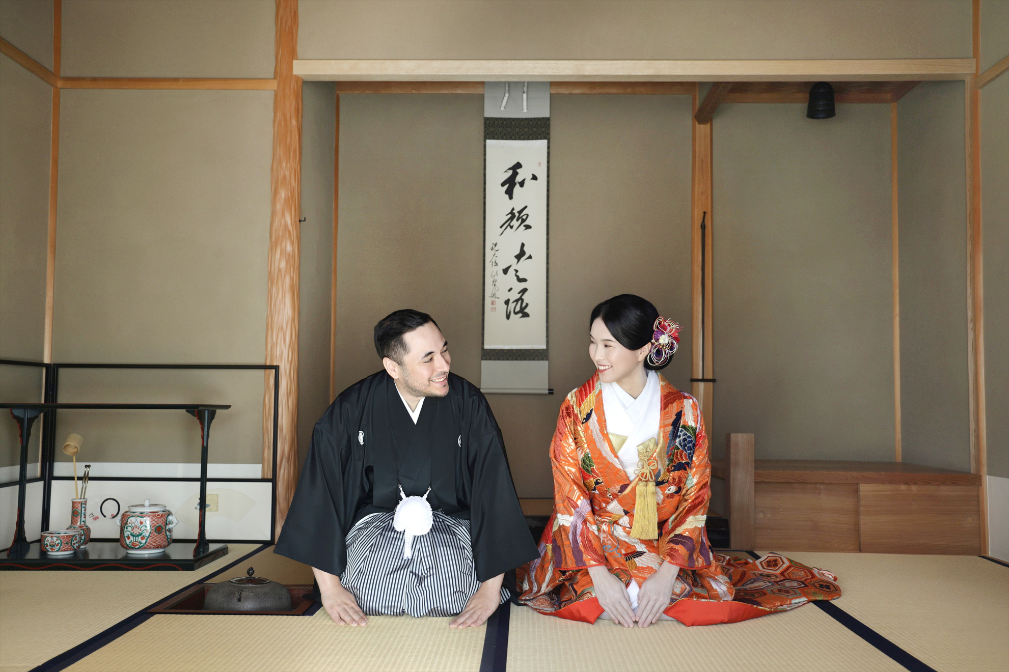 茶室の床の間を背に向かい合って笑い合う色打掛の新婦と黒紋付袴の新郎