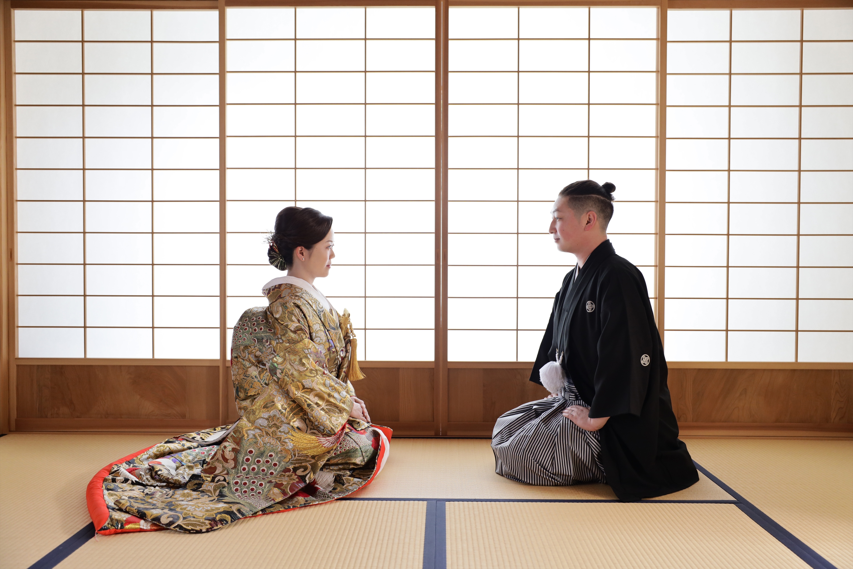 障子の光が満ちる和室で向かい合って正座する金の色打掛の新婦と黒紋付袴の新郎