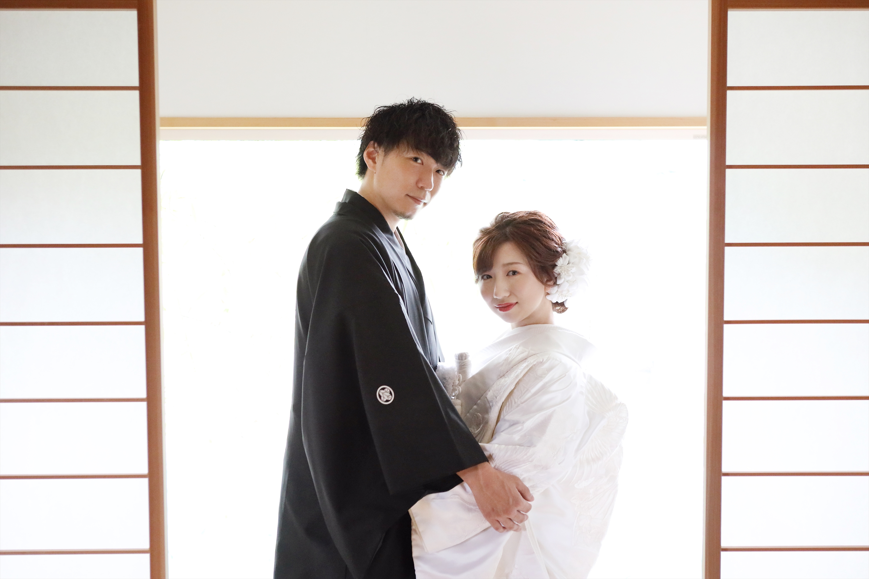 茶室の明るい障子を背に寄り添う白無垢姿の新婦と黒紋付袴の新郎
