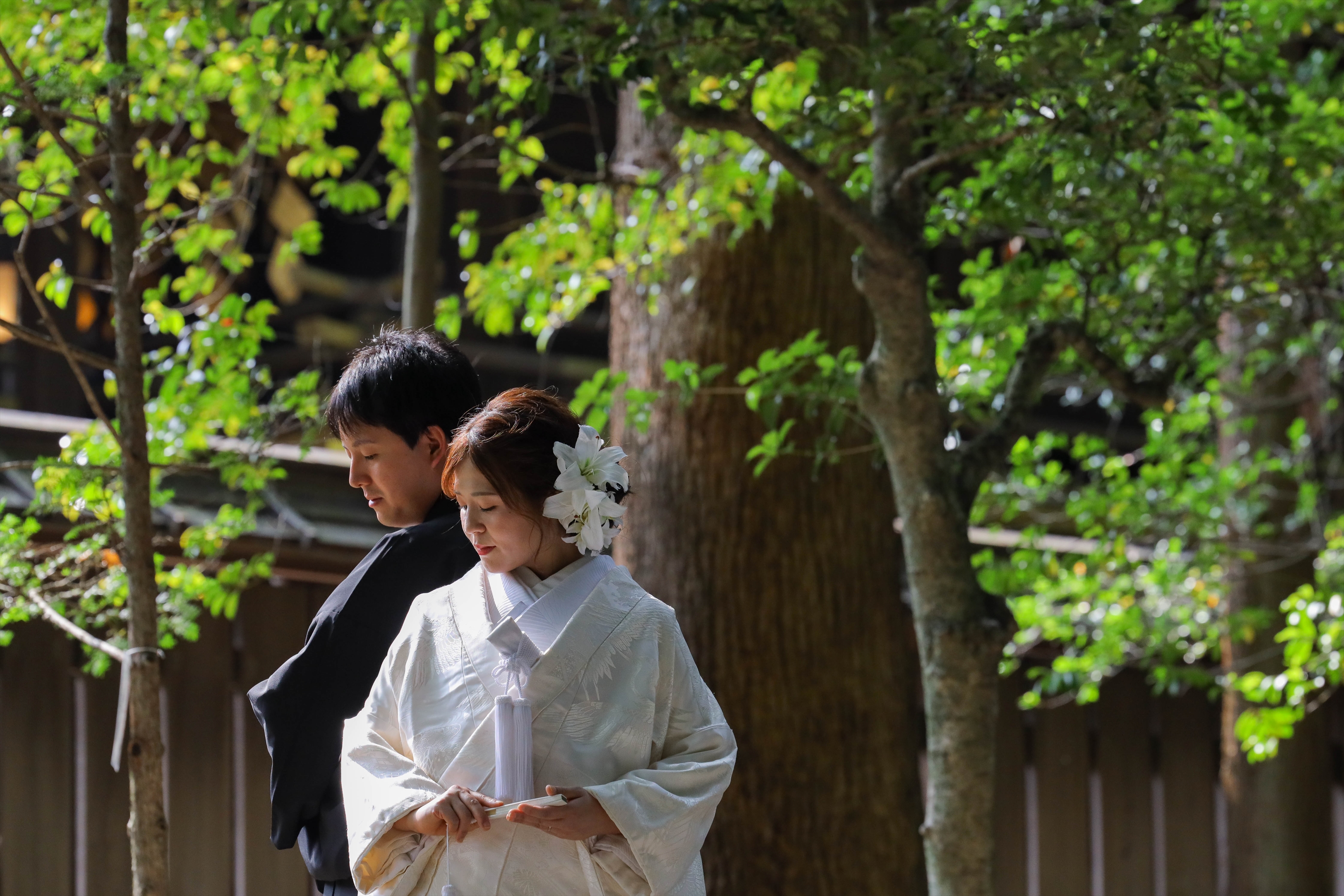 新緑の木漏れ日の下で寄り添う白百合の髪飾りに白無垢の新婦と紋付袴の新郎
