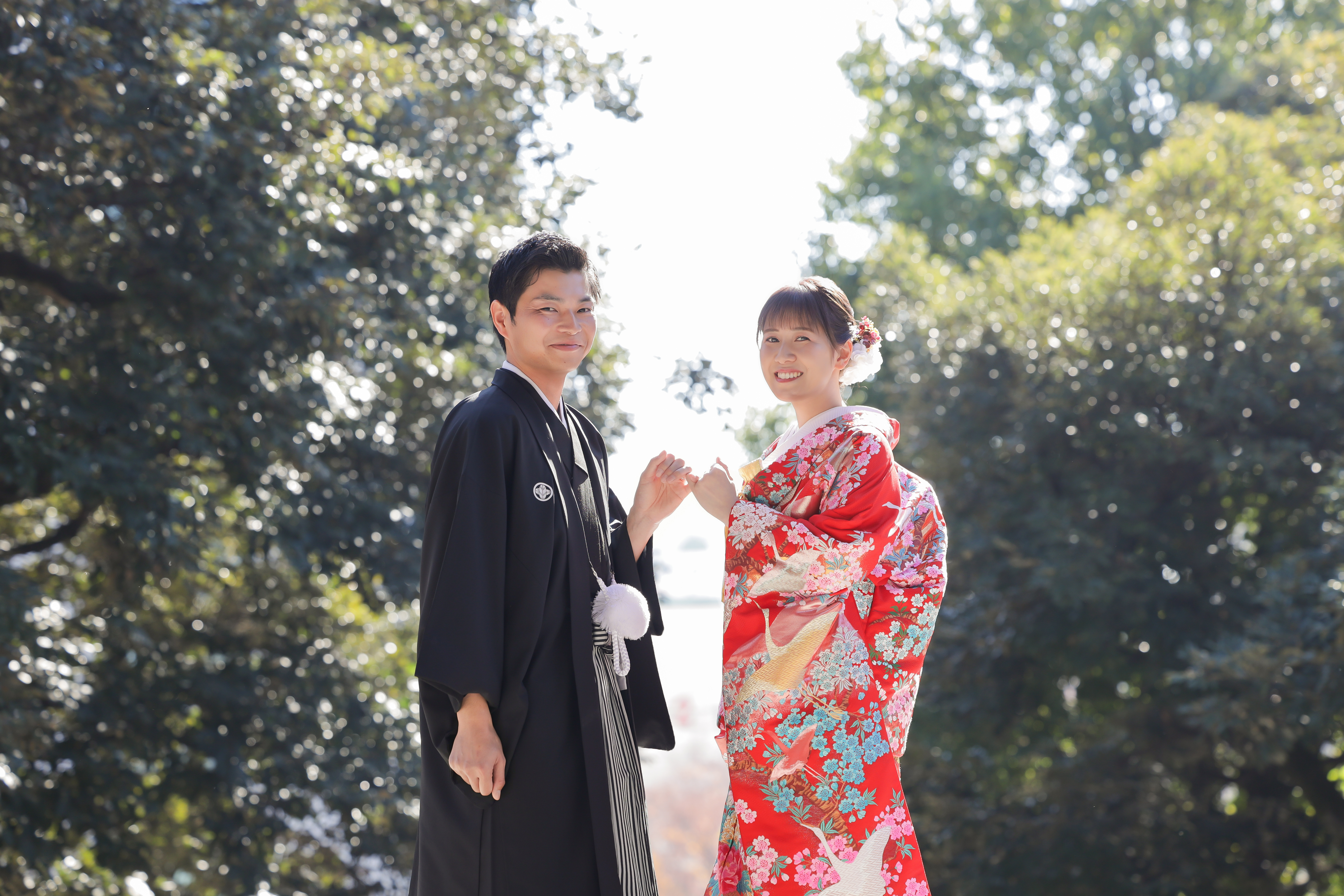 木漏れ日きらめく境内で指を絡ませ微笑む赤い色打掛の新婦と紋付袴の新郎