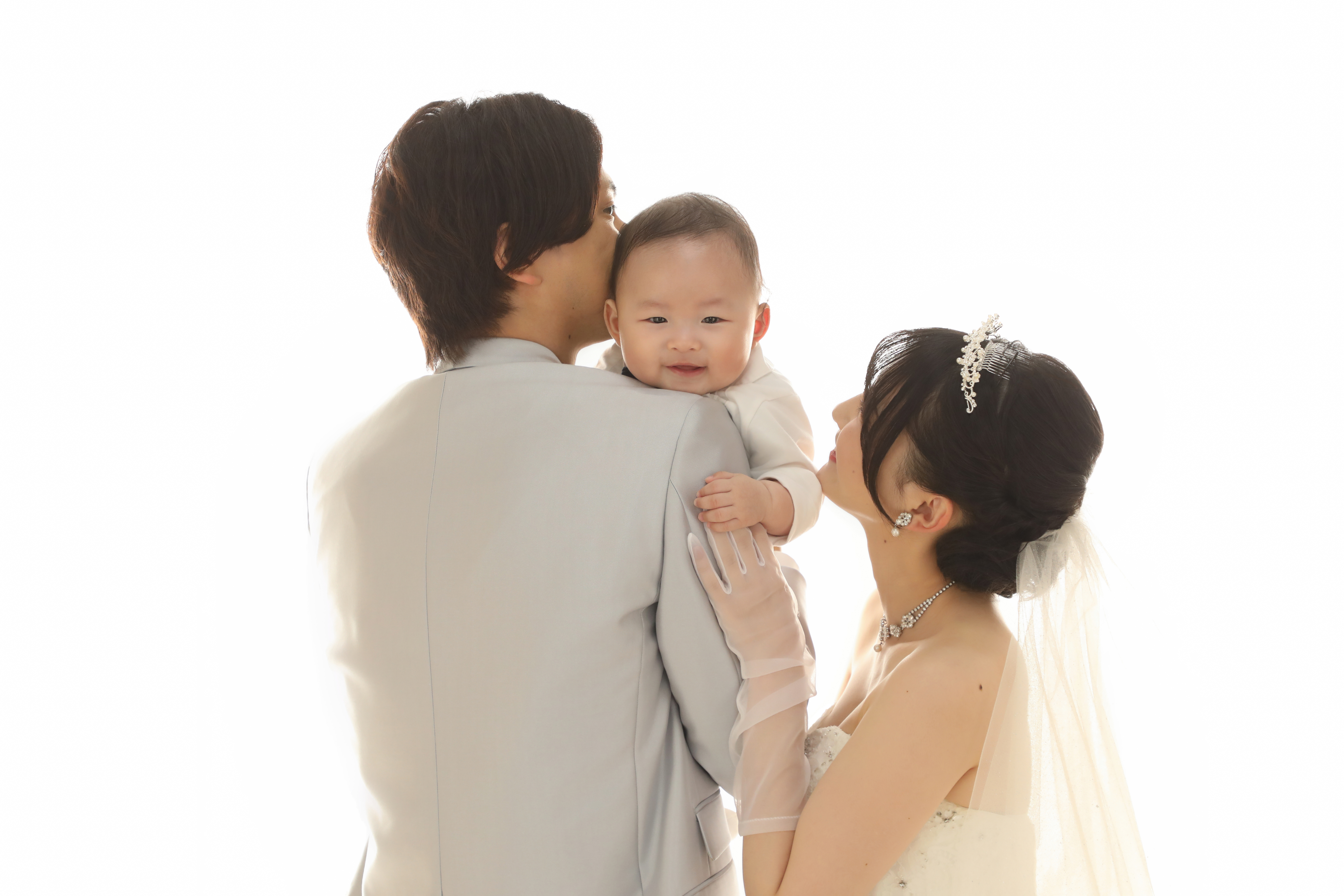 新郎の肩越しに笑う赤ちゃんを見つめるウェディングドレスの新婦