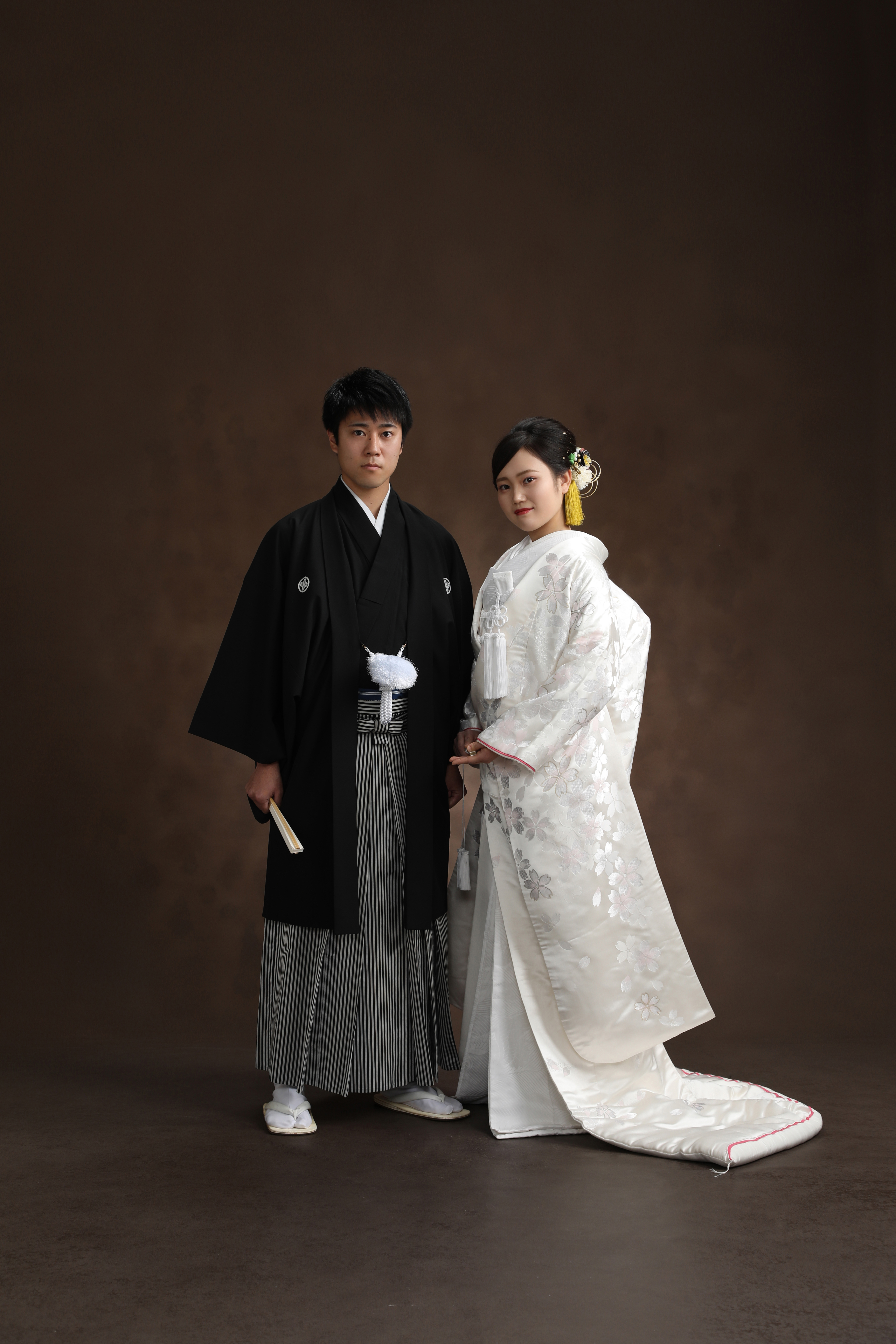 ブラウン背景で桜文様の白無垢をまとった新婦と黒紋付袴の新郎