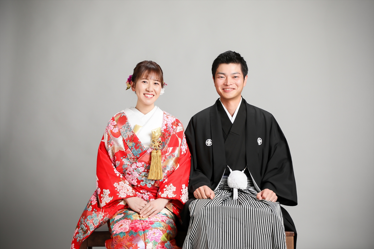 赤地に牡丹の色打掛と黒紋付袴で椅子に腰かけ微笑む新郎新婦