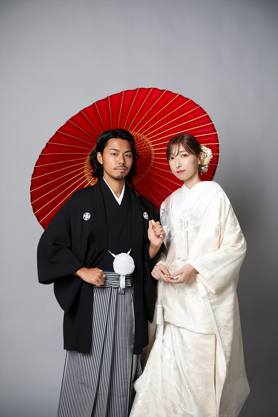 朱色の和傘を背に白無垢の新婦と黒紋付袴の新郎が寄り添う
