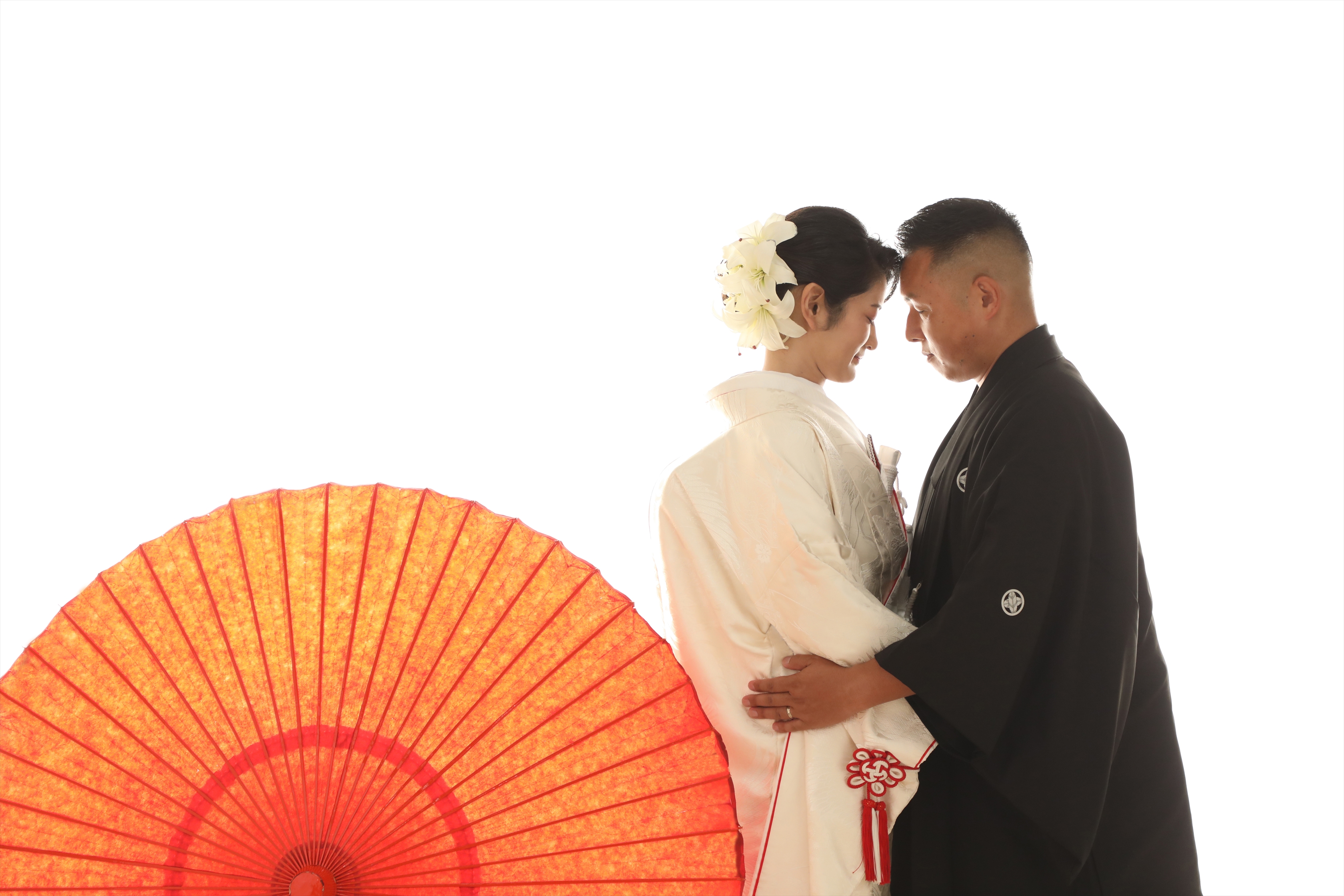 朱色の番傘のそばで額を寄せ合う白無垢の新婦と黒紋付袴の新郎