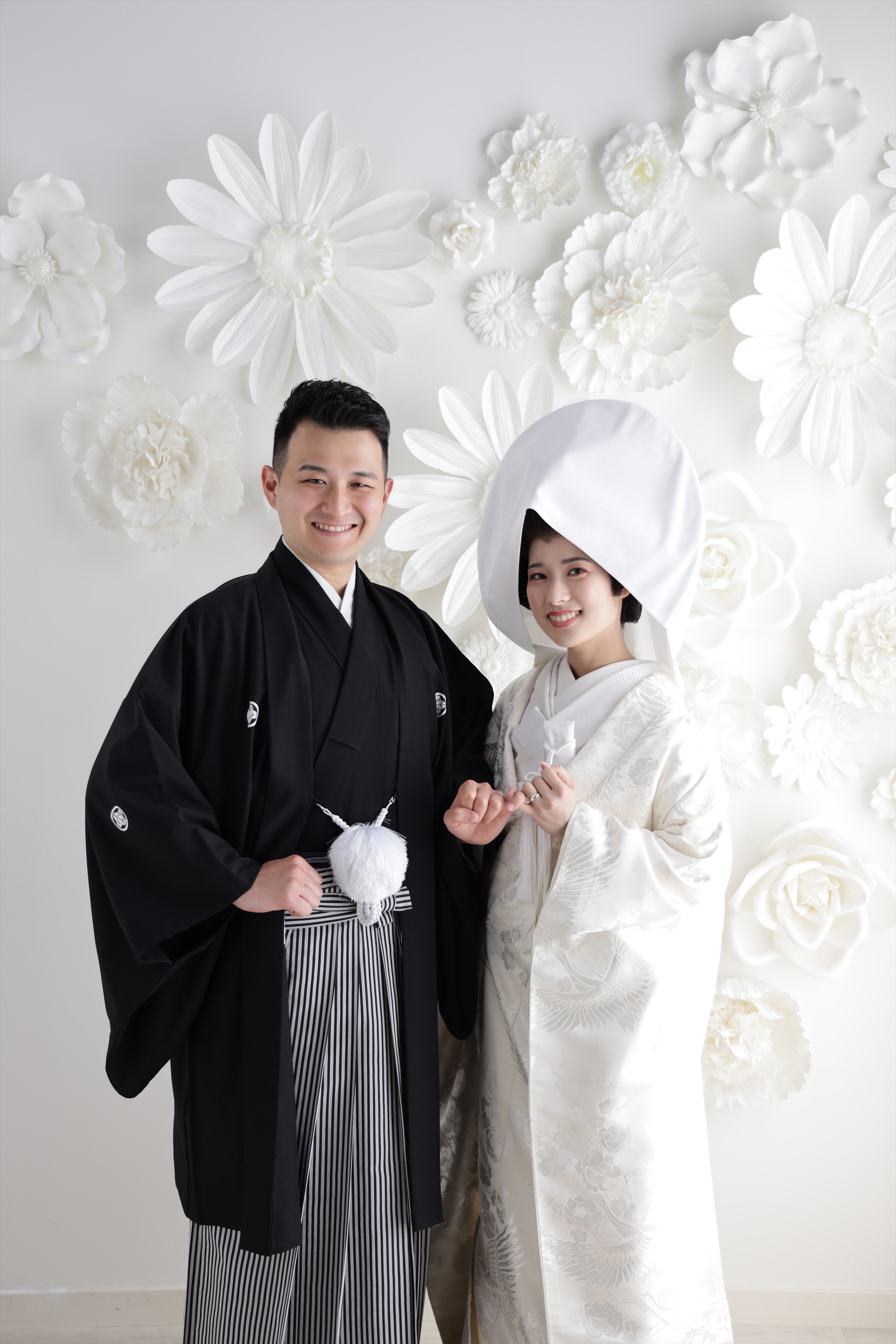 白い花の壁を背に綿帽子をかぶった白無垢の新婦と黒紋付袴の新郎