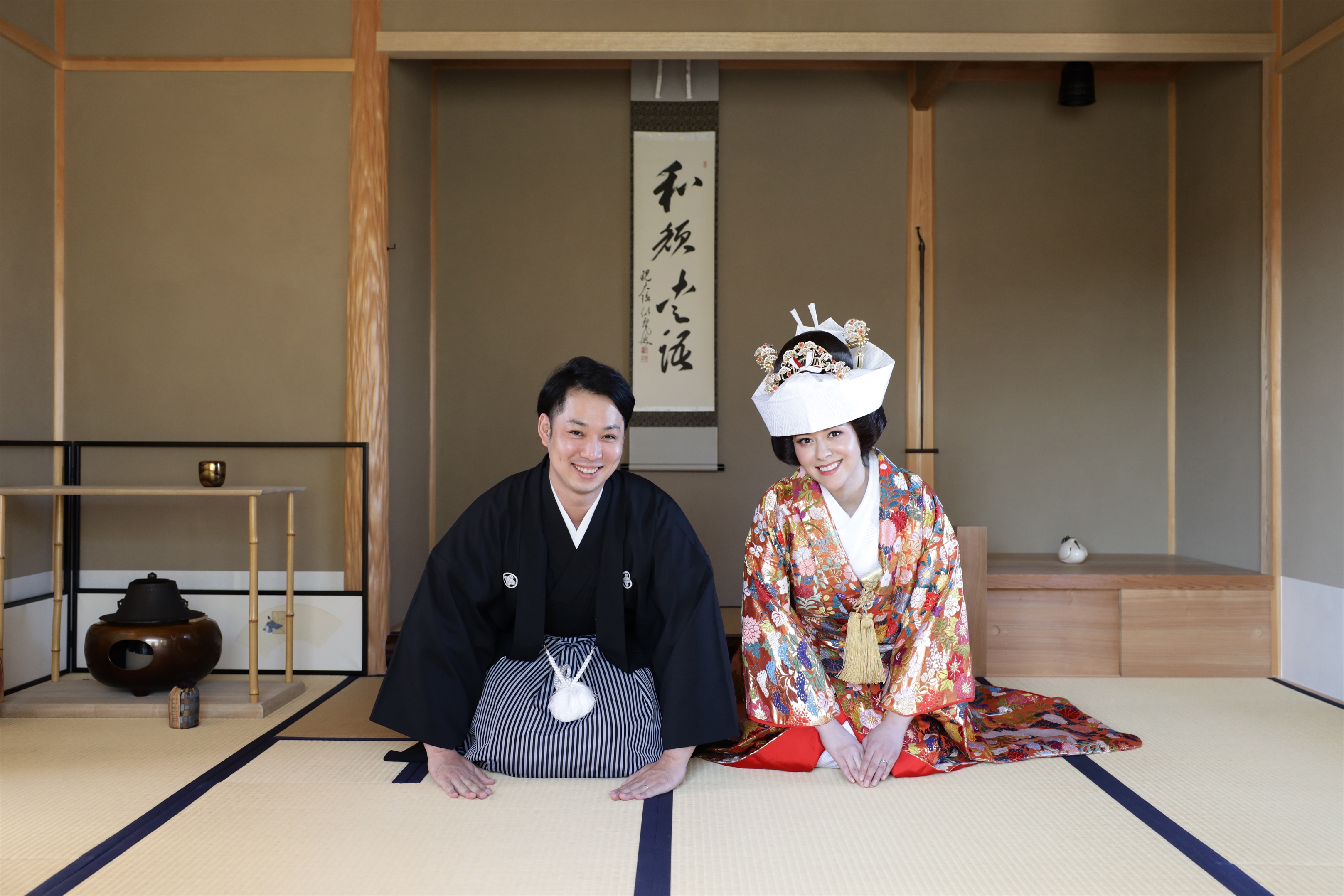 茶室の掛軸の前に正座する角隠しの色打掛の新婦と黒紋付袴の新郎