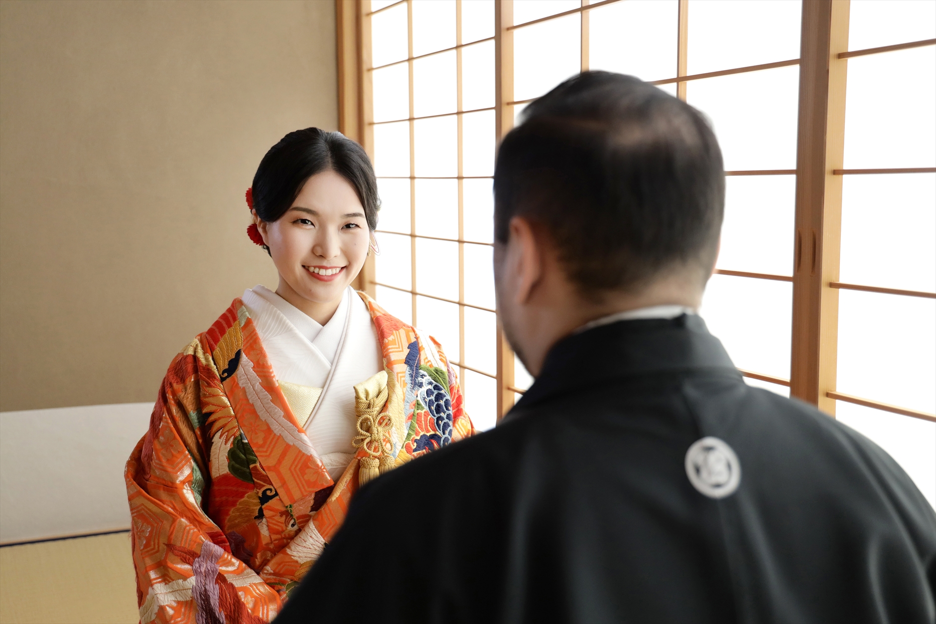 障子越しの光の中で新郎に微笑みかける朱色の色打掛の新婦