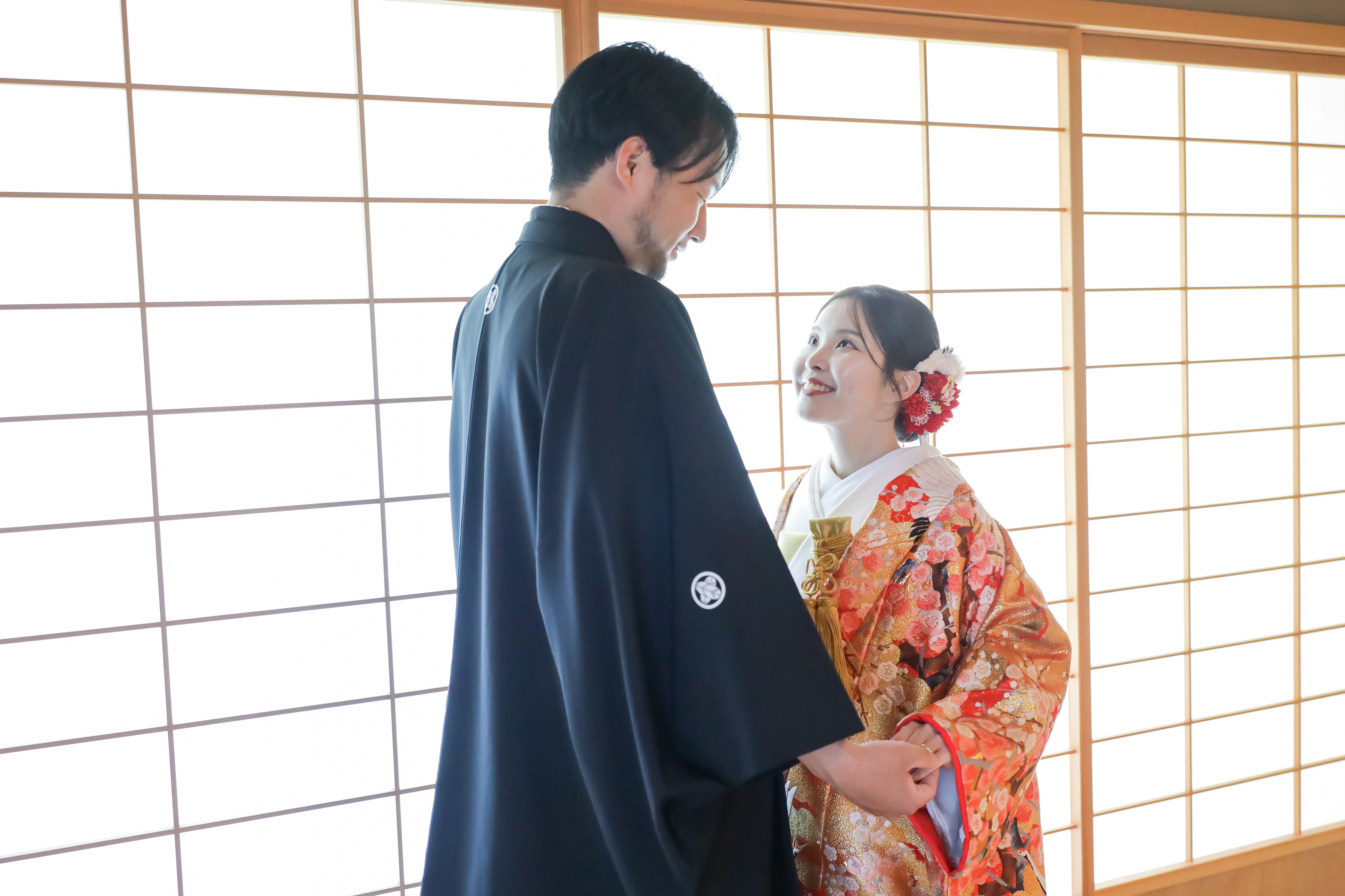 障子の光に包まれ手を取って見つめ合う梅柄の色打掛と黒紋付袴の二人