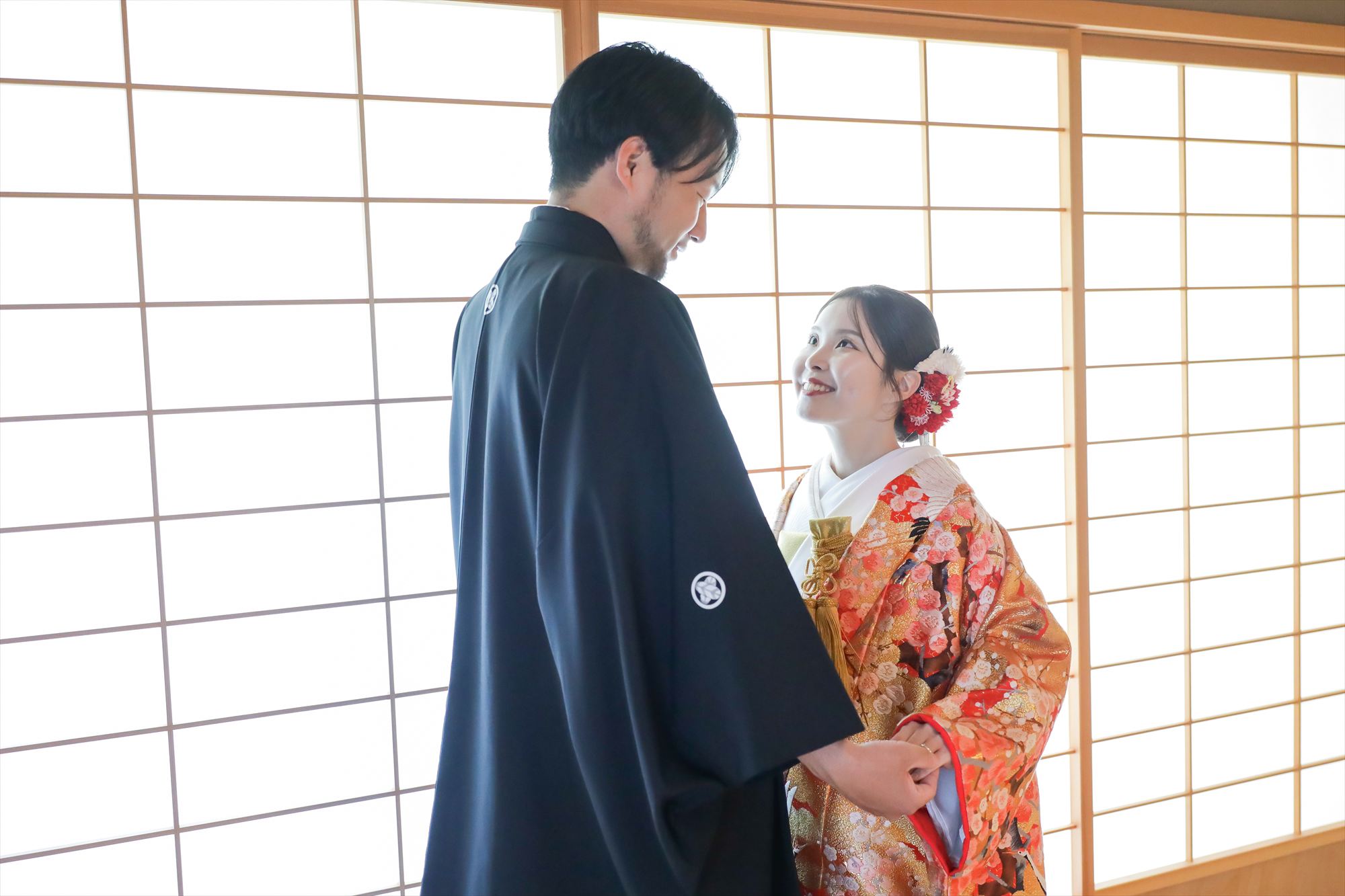 障子の光に包まれ手を取って見つめ合う梅柄の色打掛と黒紋付袴の二人