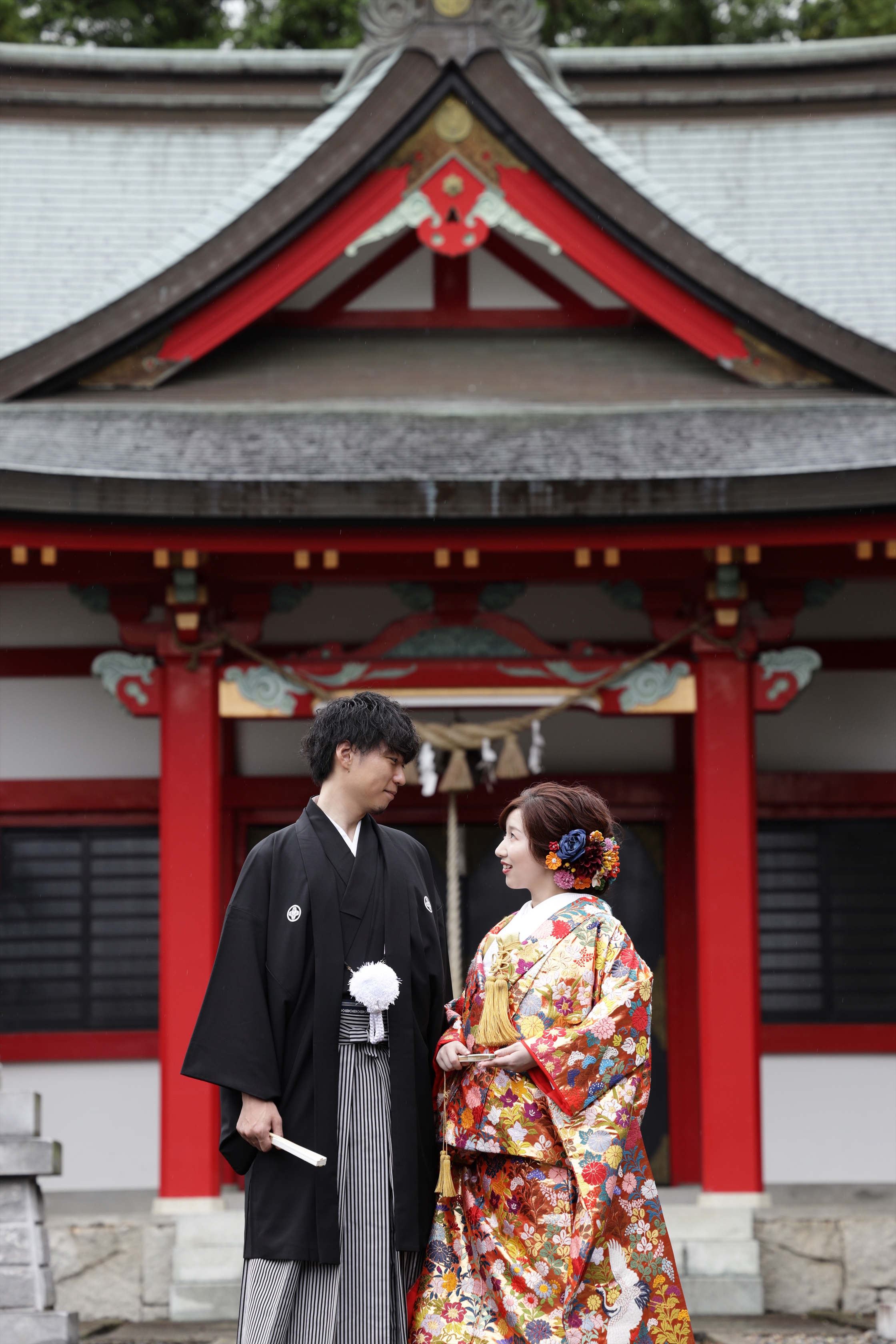 朱塗りの社殿を背に見つめ合う色打掛の新婦と黒紋付袴の新郎