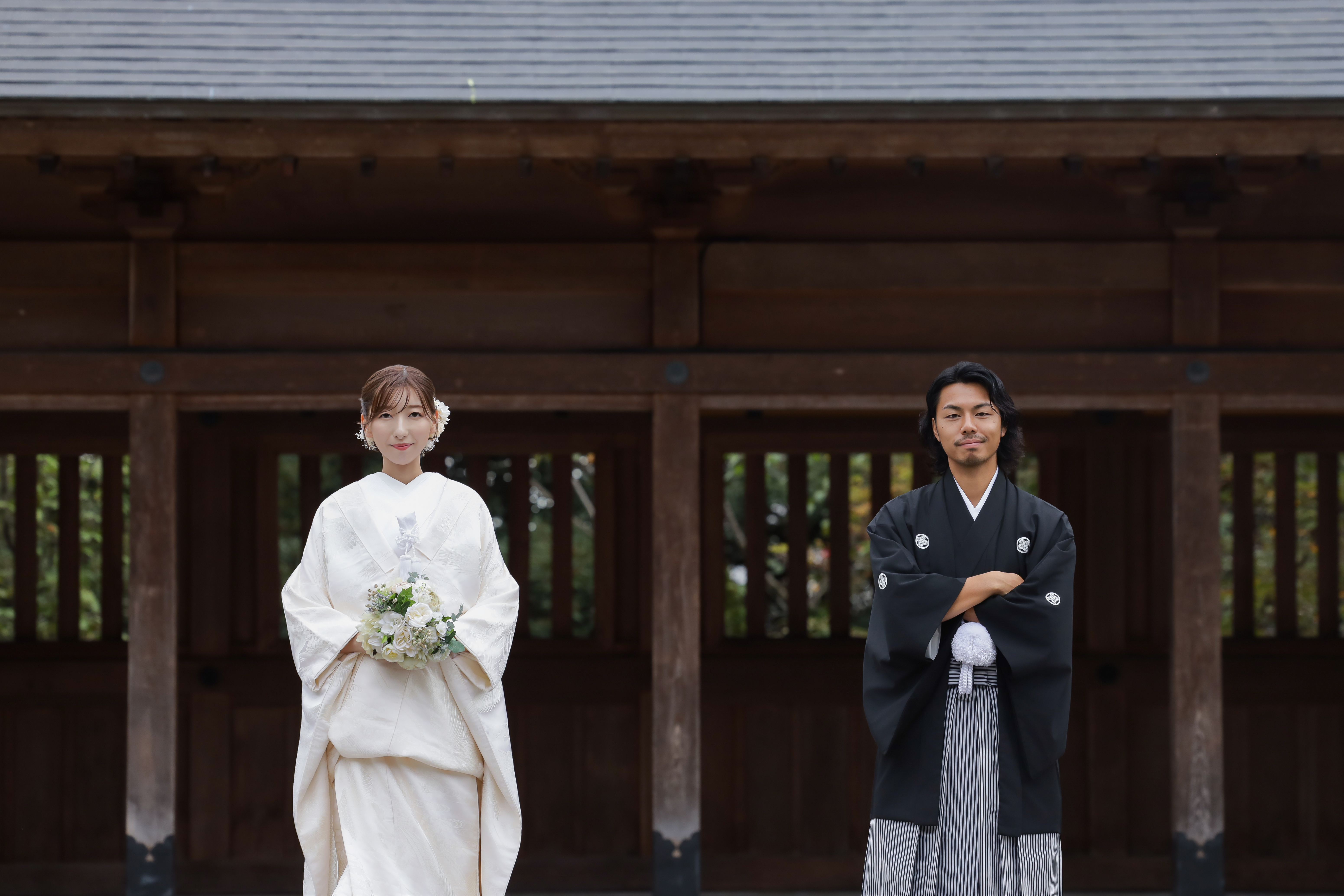 社殿を背に離れて立つブーケを持つ白無垢の新婦と腕を組む黒紋付袴の新郎
