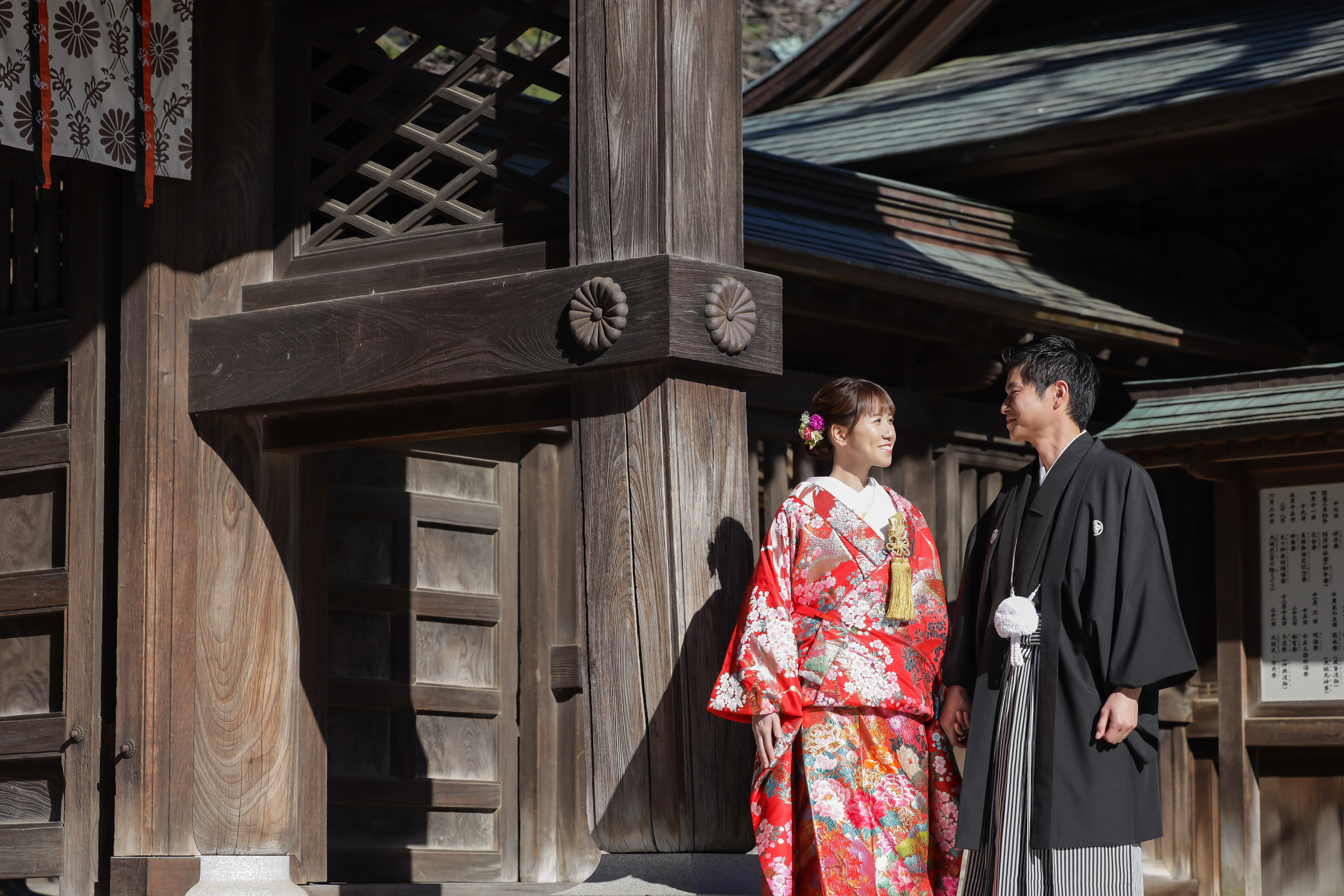 神社の大きな木柱のそばで見つめ合う赤い色打掛の新婦と黒紋付袴の新郎