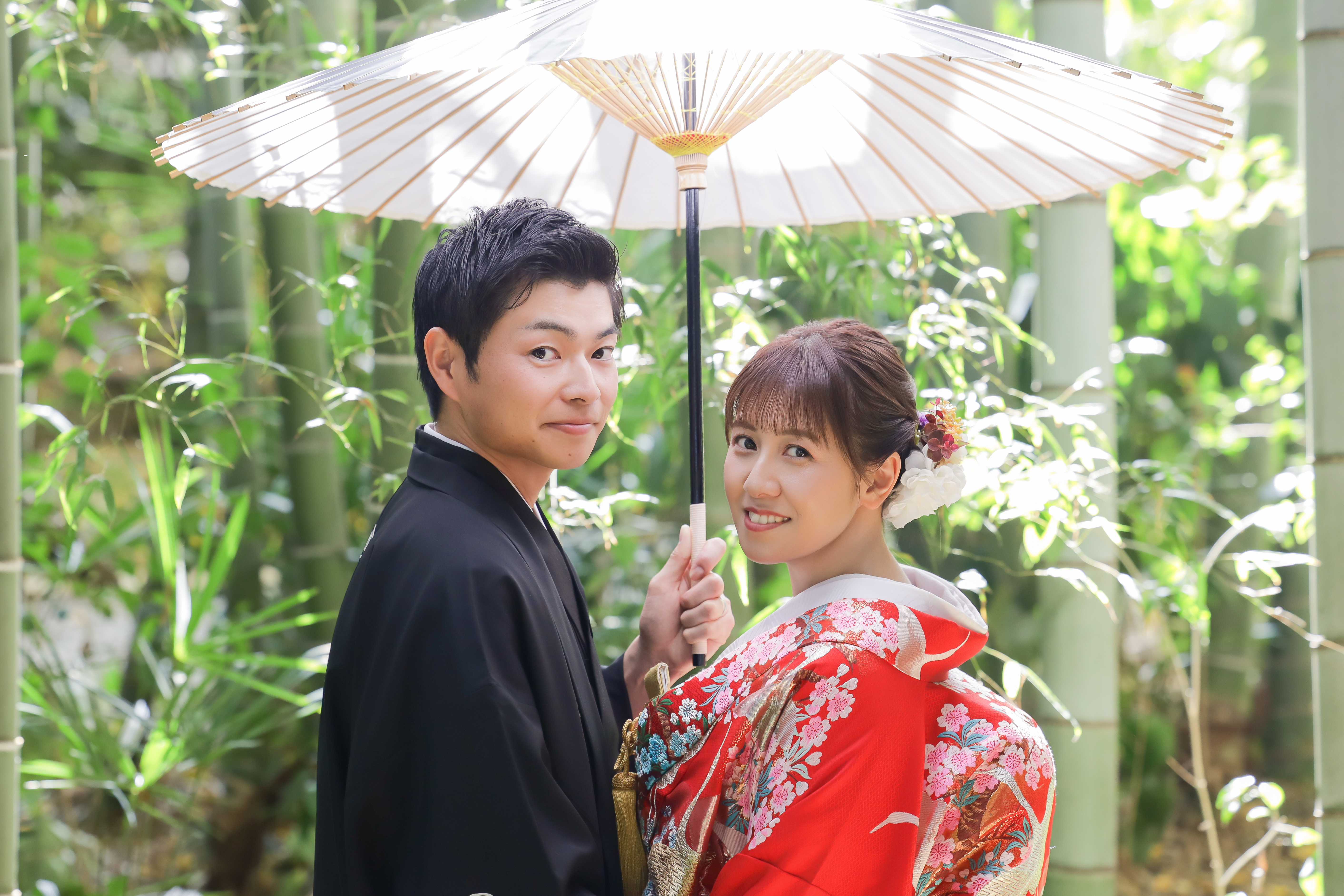 竹林で白い和傘を差して振り返る赤い色打掛の新婦と黒紋付袴の新郎