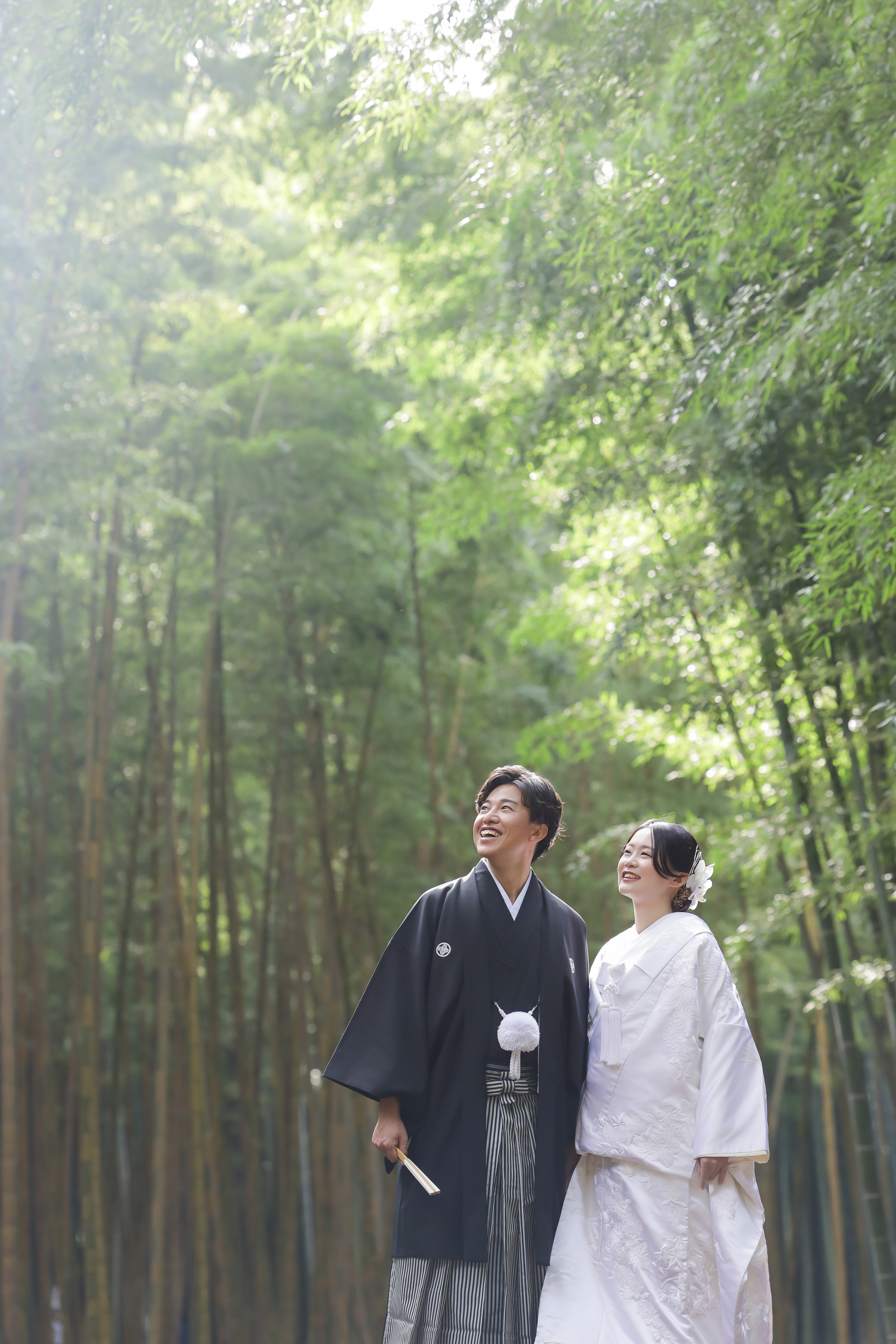 竹林の光を見上げて笑い合う白無垢の新婦と黒紋付袴の新郎