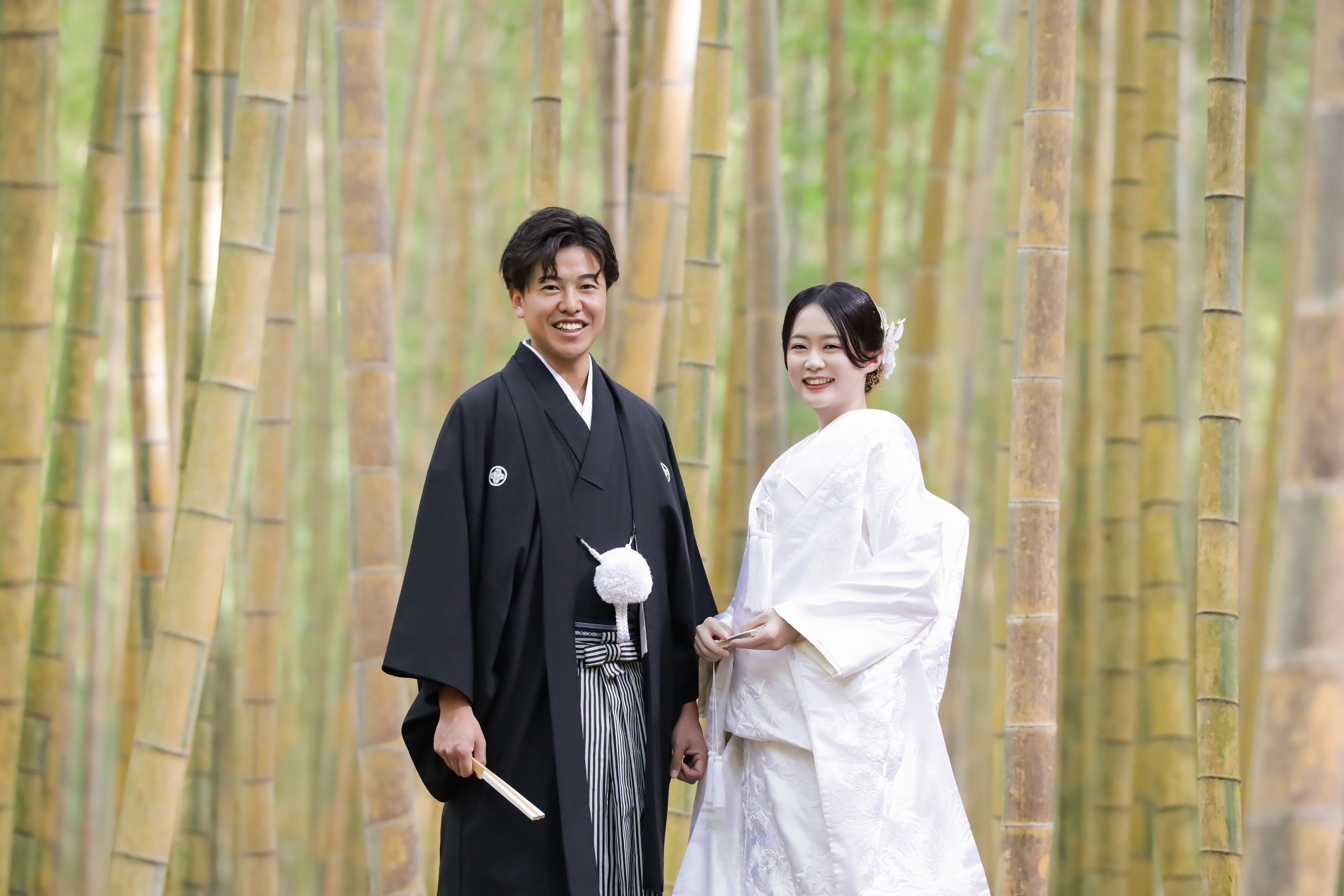 竹林の中で扇子を手に並んで微笑む白無垢の新婦と黒紋付袴の新郎