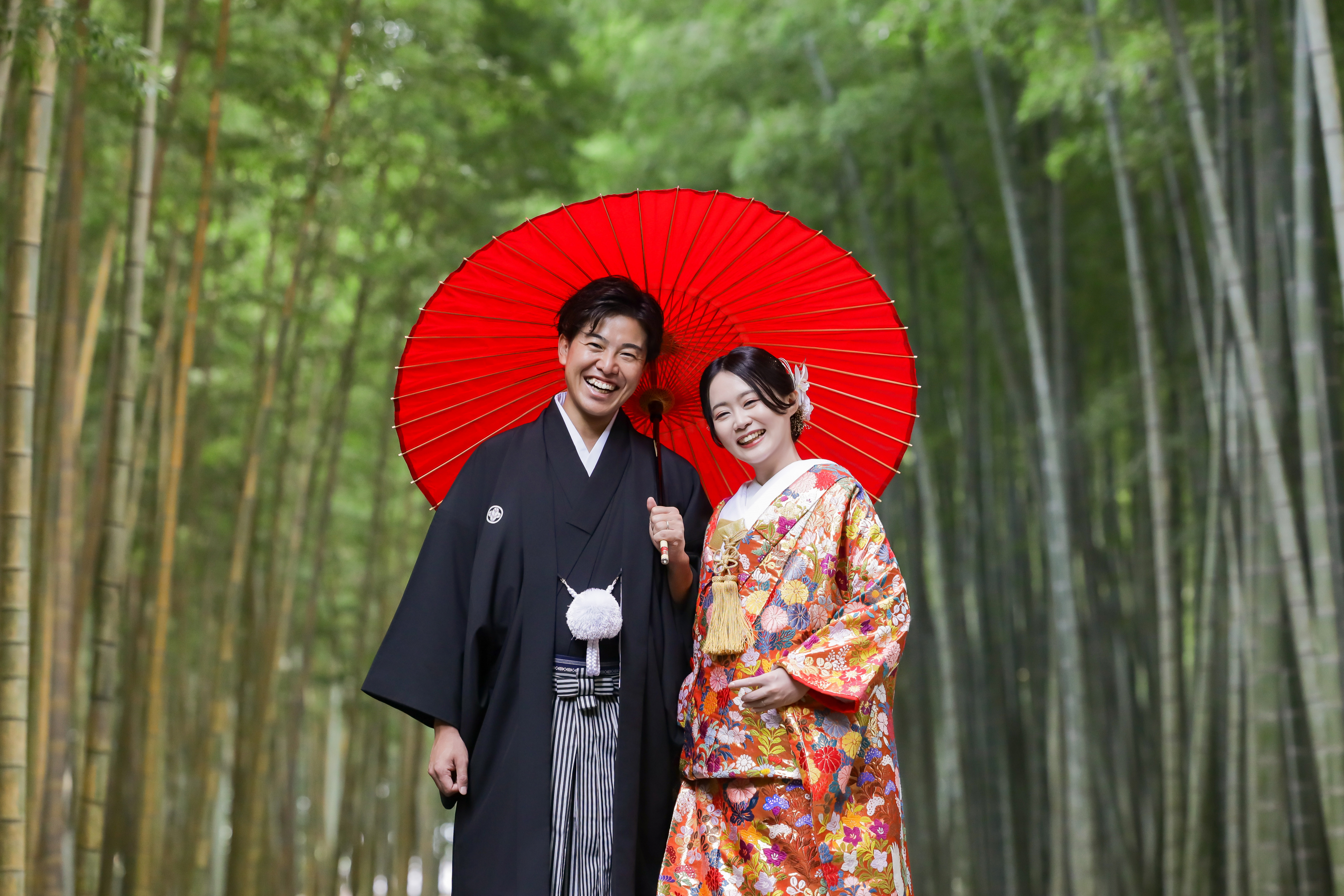 竹林で赤い和傘を掲げて笑顔を見せる色打掛の新婦と黒紋付袴の新郎