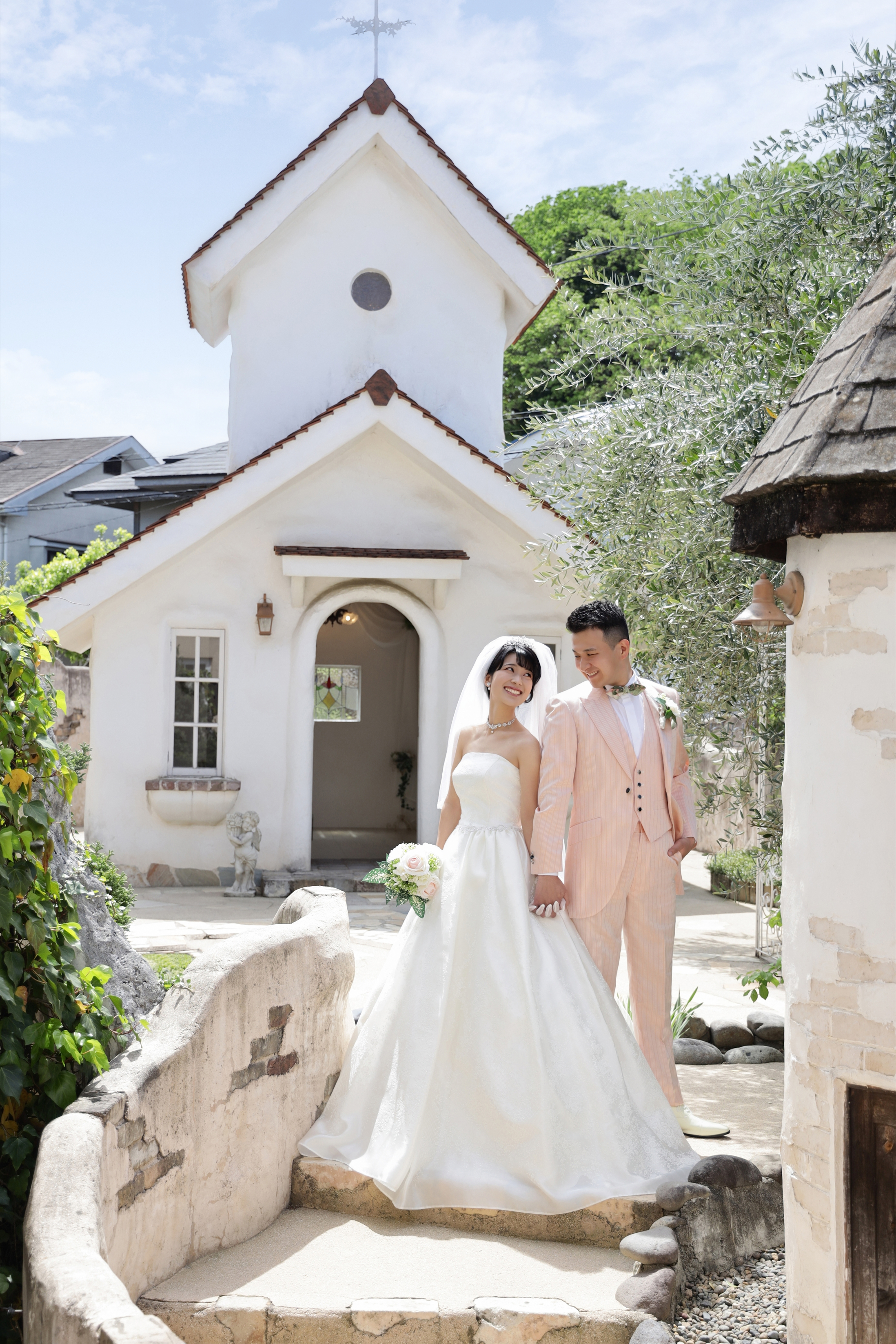 白いチャペルの前で寄り添うウェディングドレスの新婦とピンクのタキシードの新郎