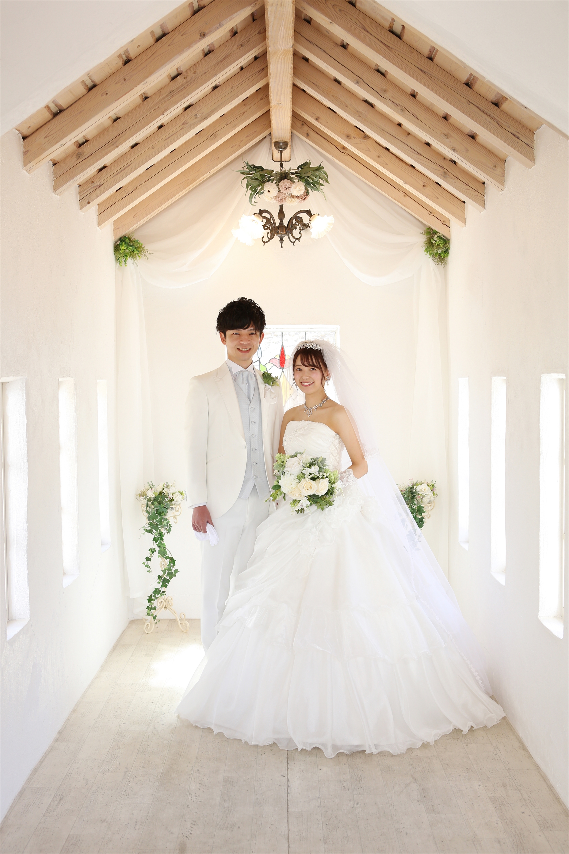 白い漆喰のチャペル内でウェディングドレスと白タキシードで並ぶ新郎新婦