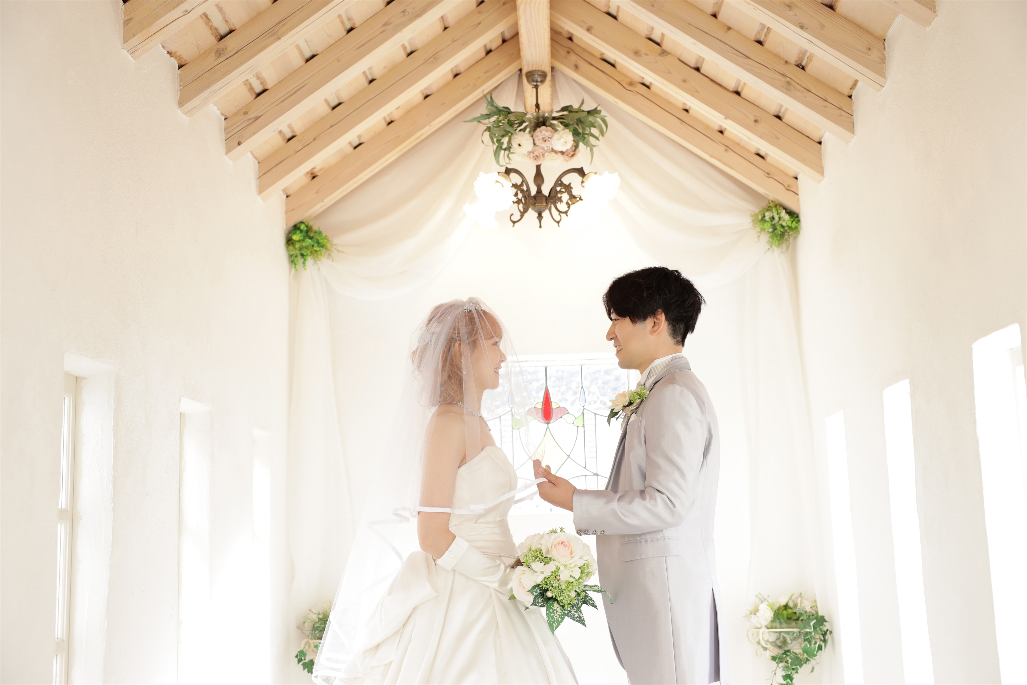光あふれるチャペルでベール越しに見つめ合うドレスの新婦とグレータキシードの新郎