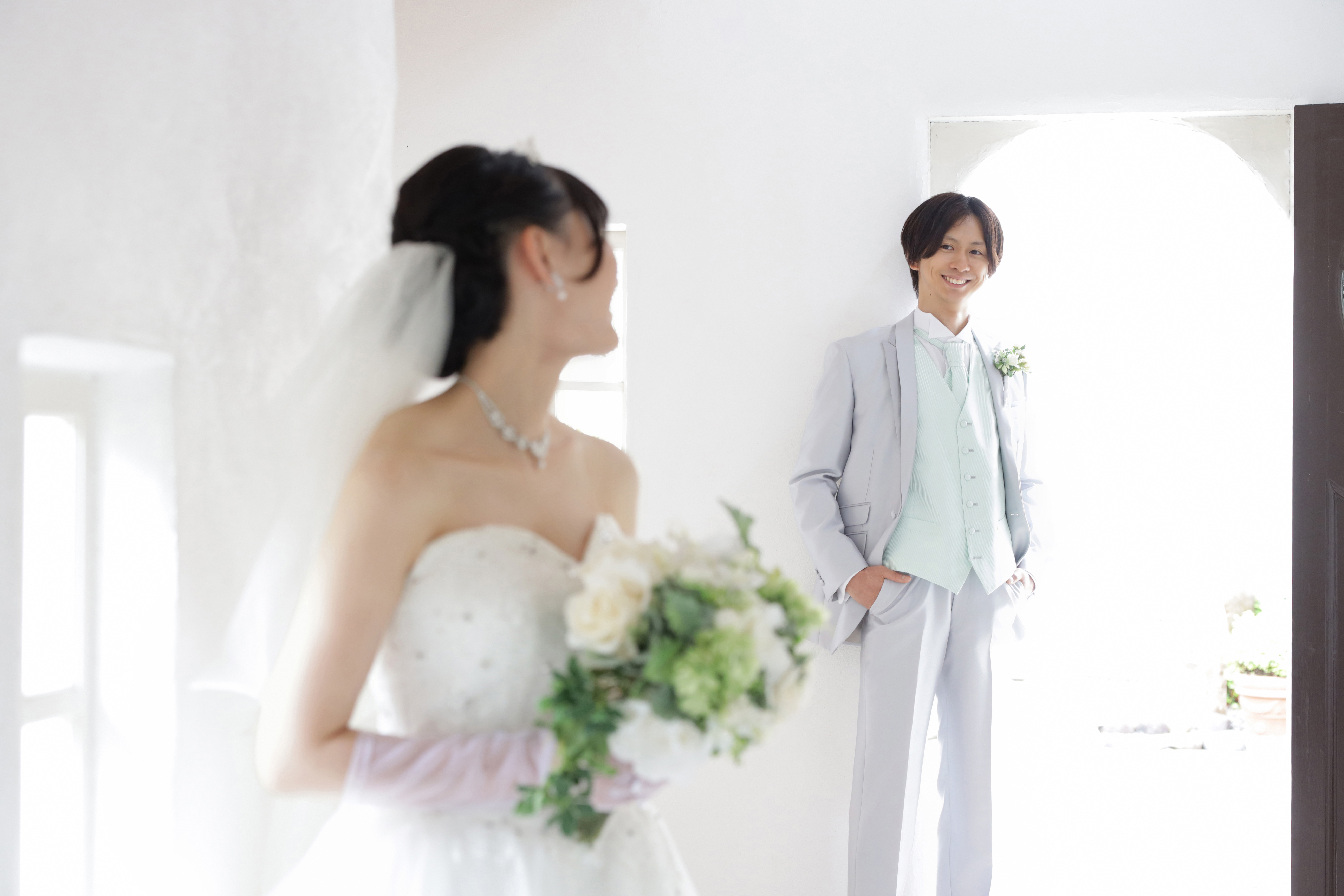白い室内でブーケを持つ新婦と壁際にたたずむグレータキシードの新郎