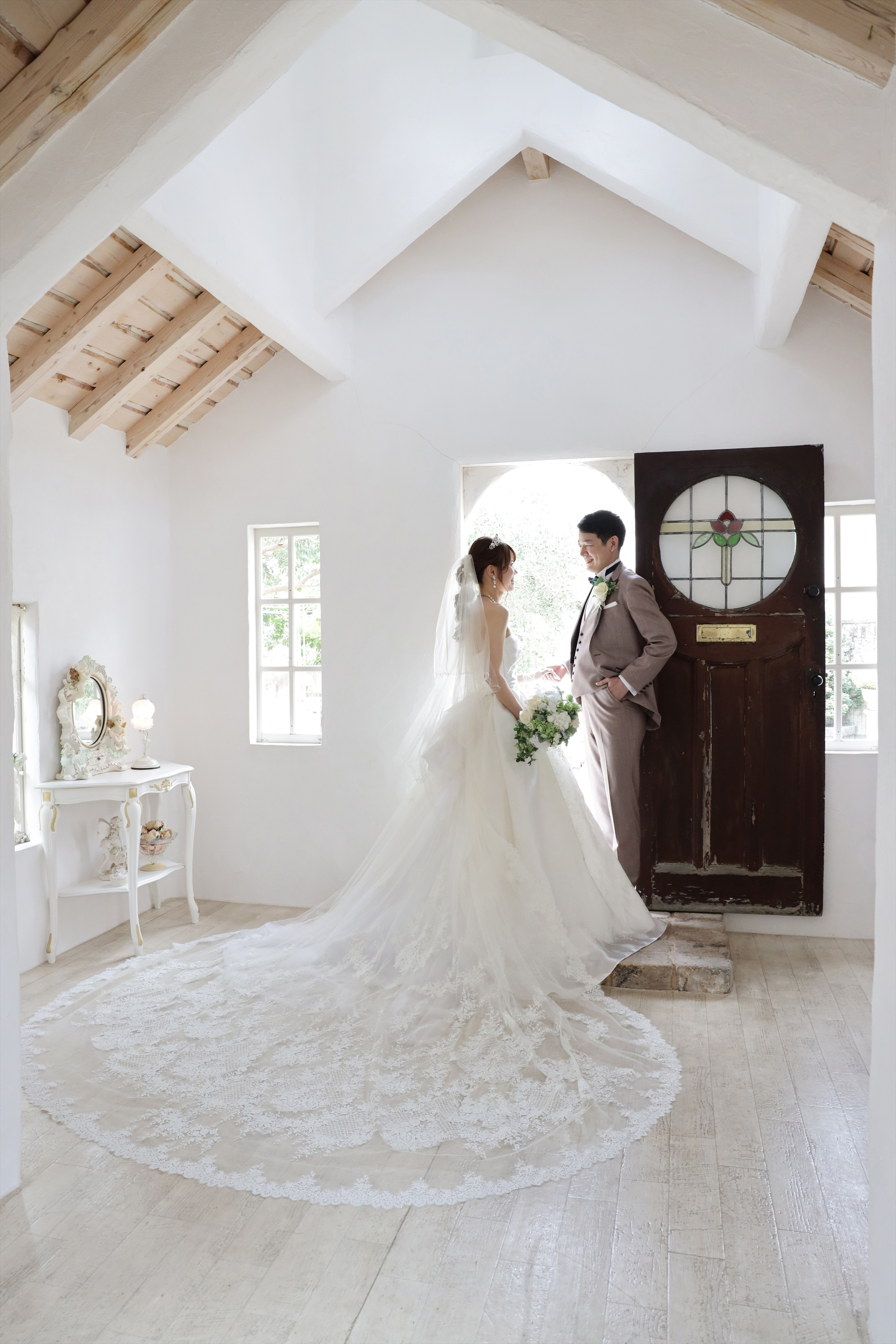 長いレースのトレーンを広げたウェディングドレスの新婦とアンティーク扉前の新郎