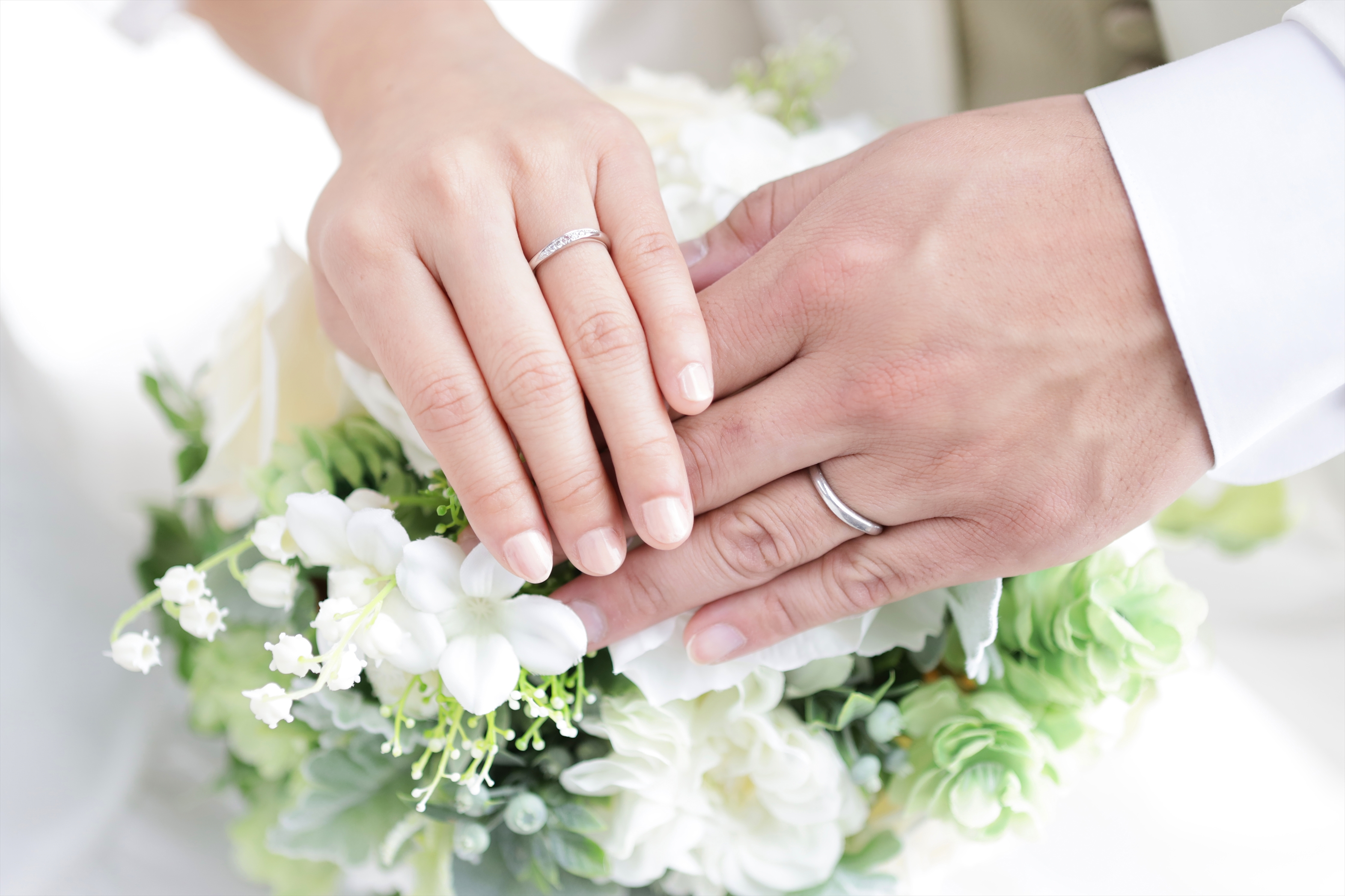 白いブーケの上に重ねた結婚指輪をつけた新郎新婦の手元