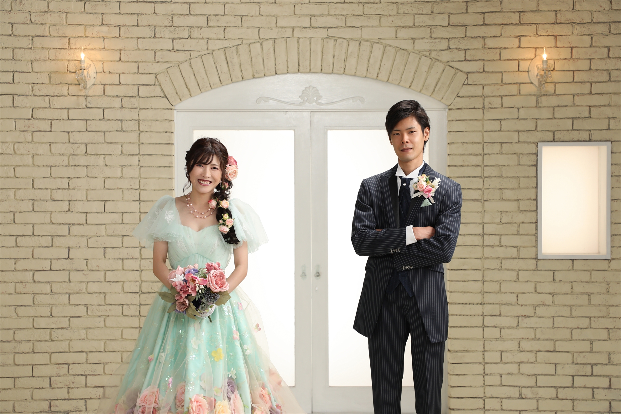 レンガ壁を背にミントグリーンのカラードレスの新婦とストライプ礼服の新郎