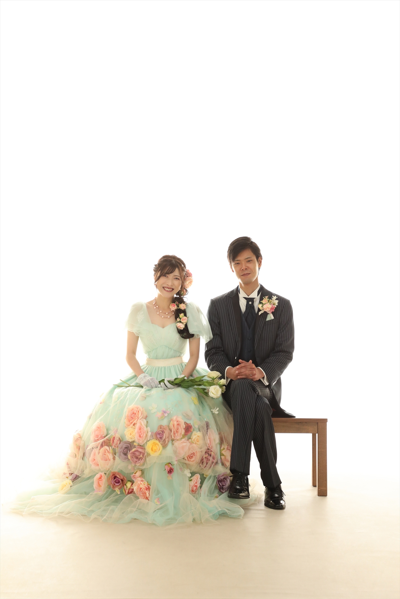 白ホリゾントでベンチに腰かけたミントグリーンの花柄ドレスの新婦と礼服の新郎