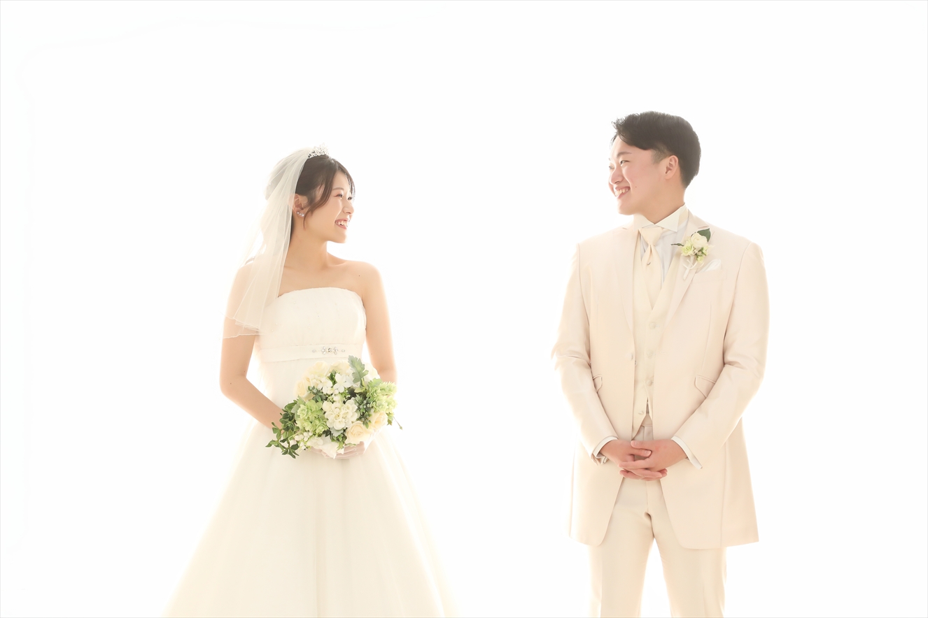 ハイキーな白背景で見つめ合うウェディングドレスの新婦とアイボリータキシードの新郎