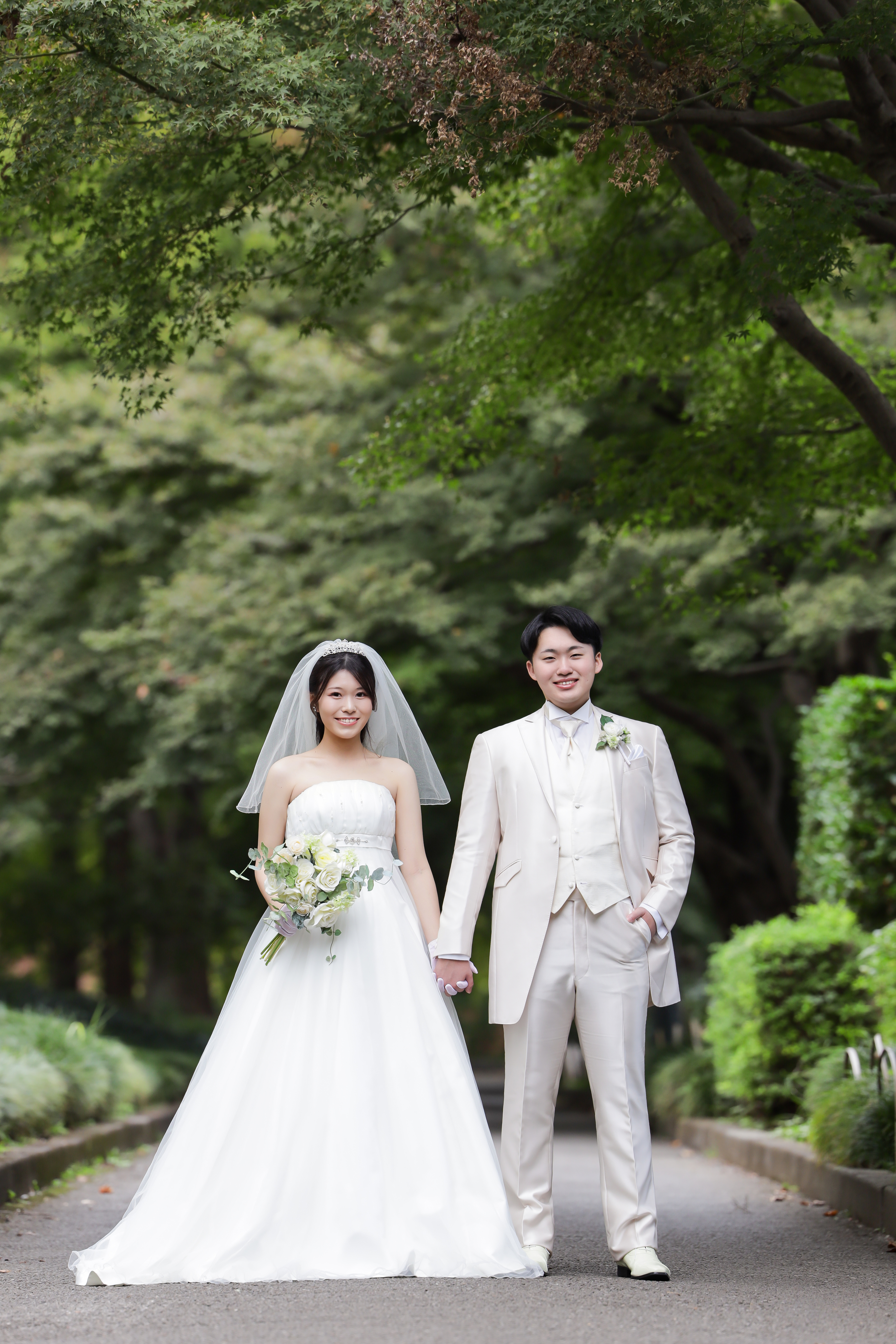 新緑の並木道で手をつなぐウェディングドレスの新婦と白タキシードの新郎
