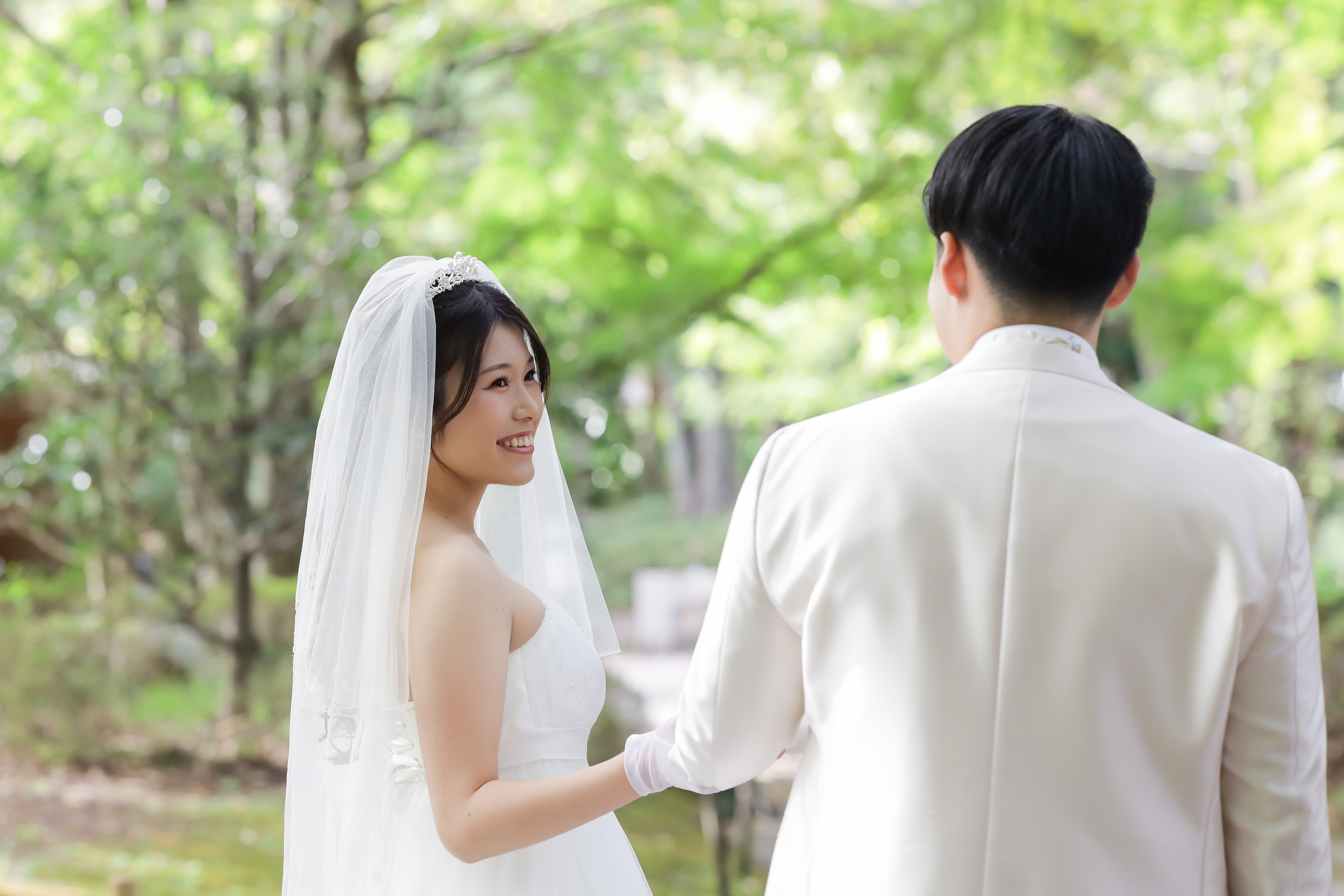 木漏れ日の中で新郎に手を引かれ振り返って微笑むベール姿の新婦
