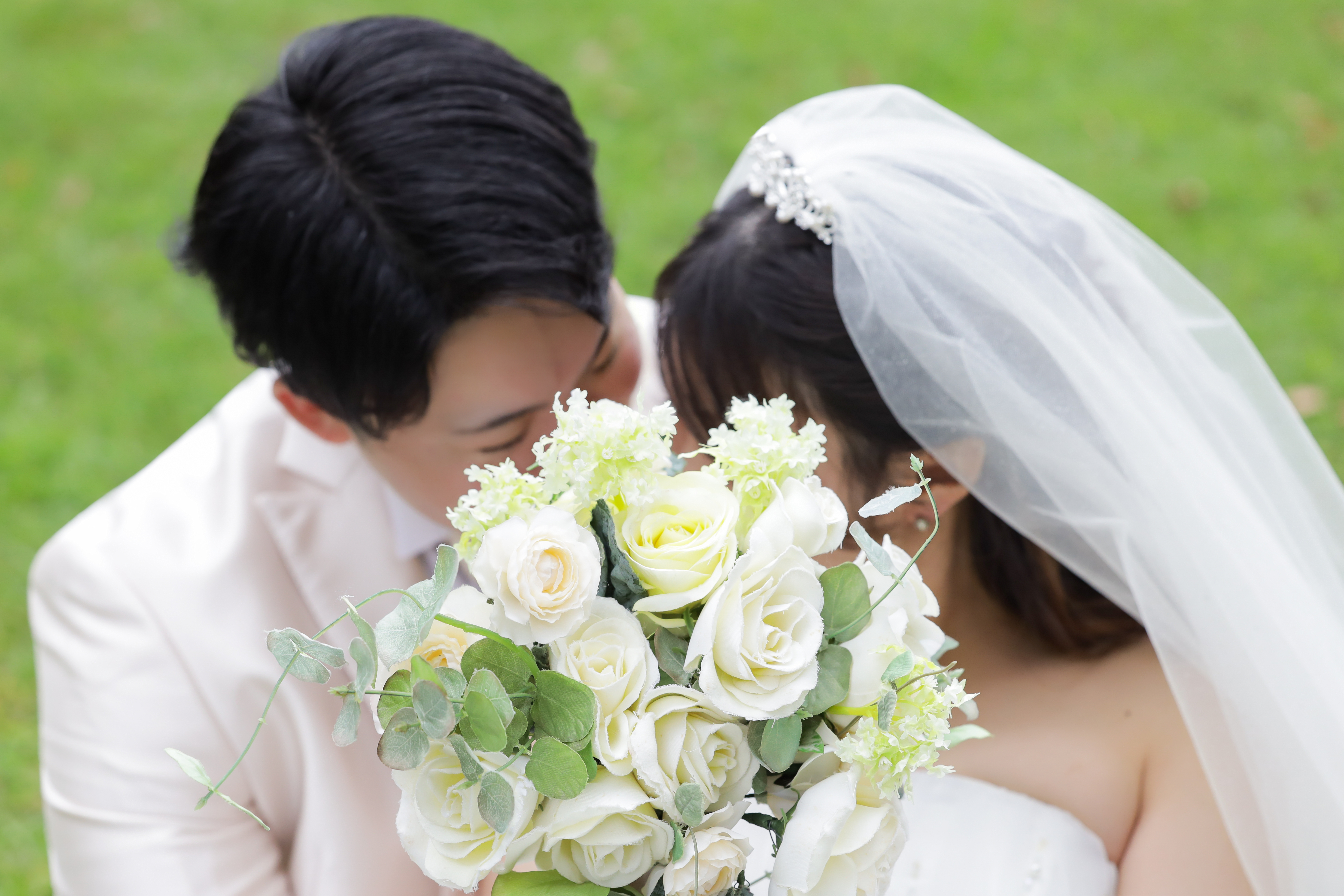 芝生の上で白いバラのブーケ越しに寄り添うベールの新婦と白タキシードの新郎