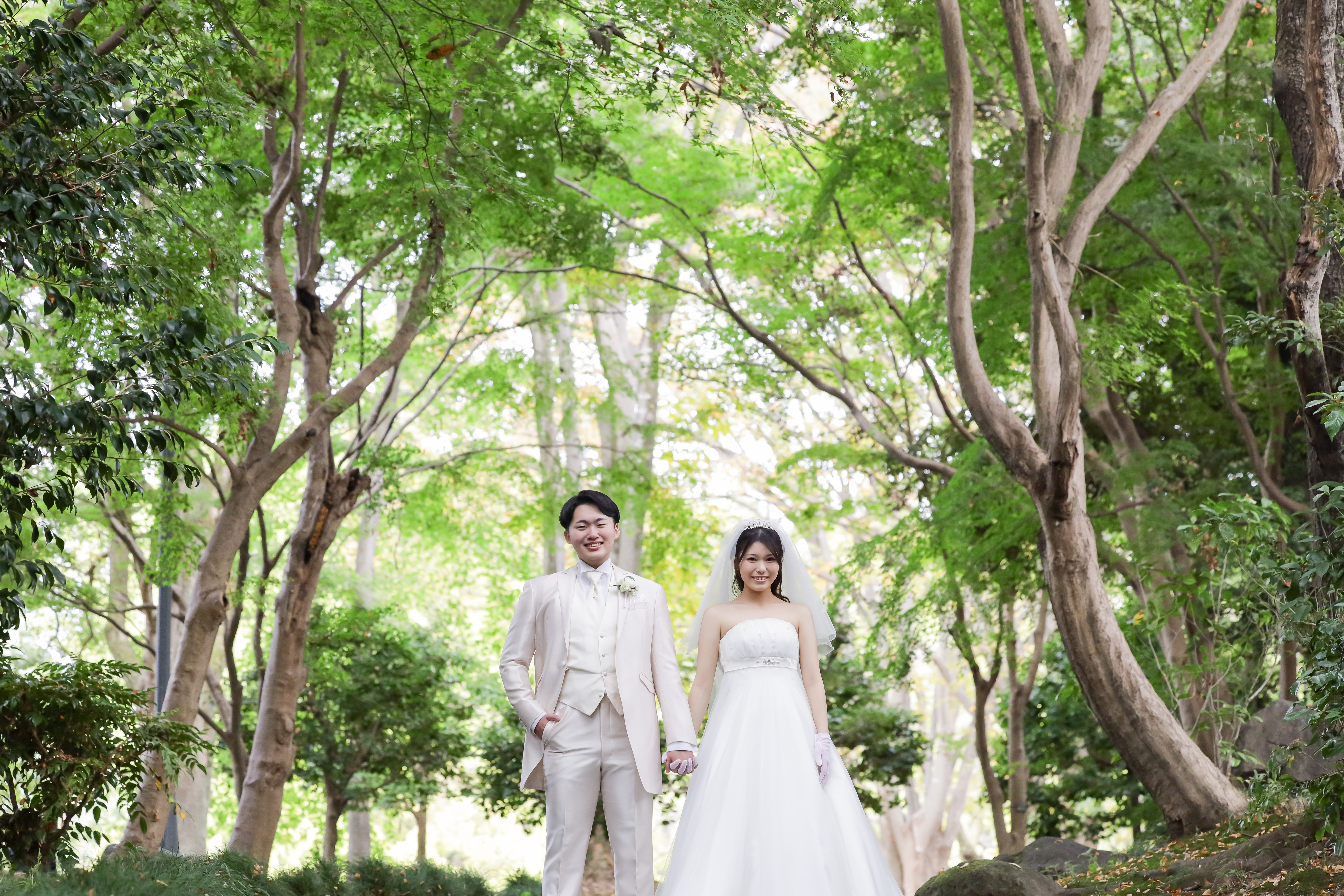 新緑の公園で手をつないで並ぶ白タキシードの新郎とベールの新婦