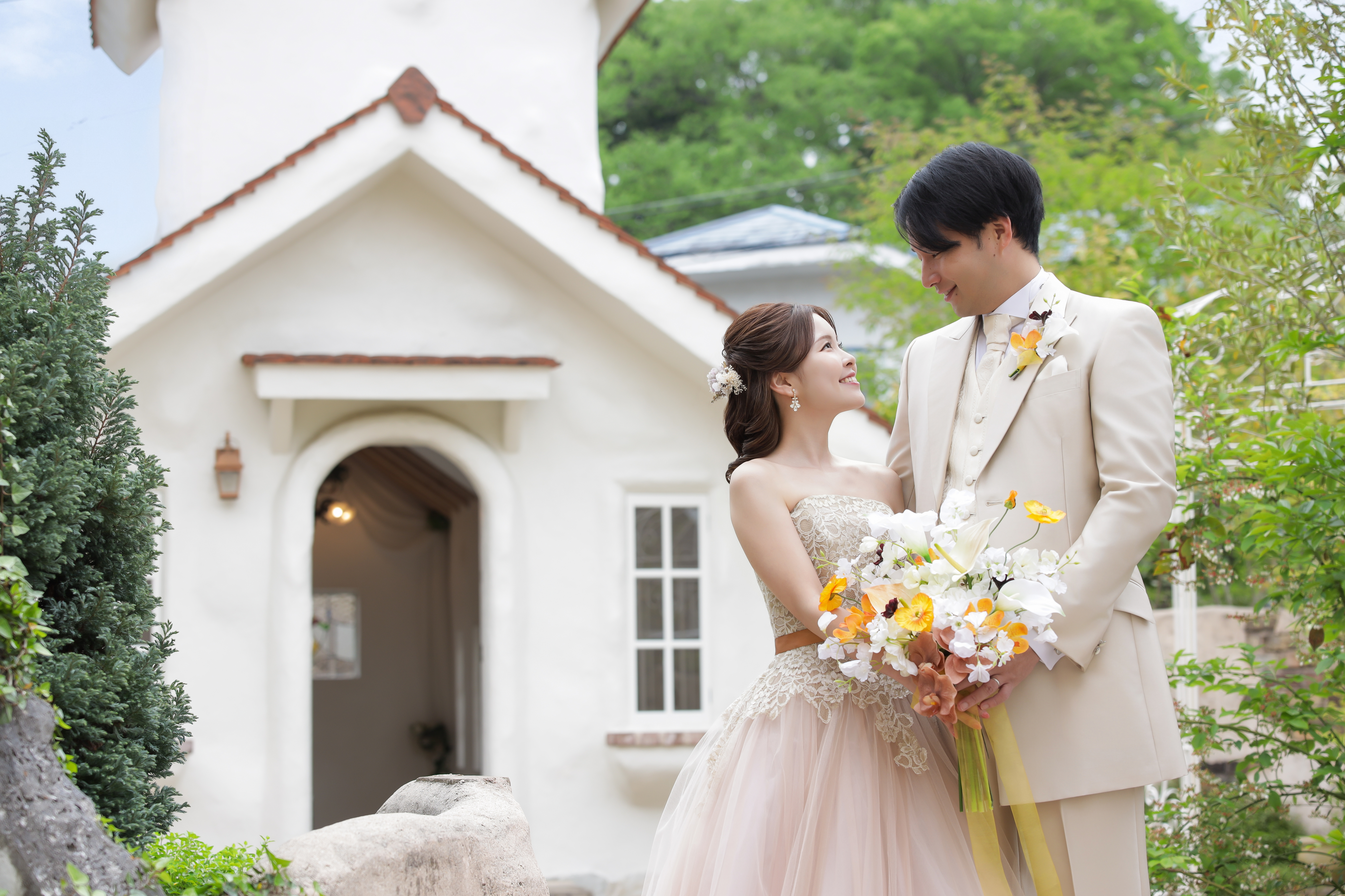 白い教会の前で見つめ合うベージュのウェディングドレスの新婦とタキシードの新郎