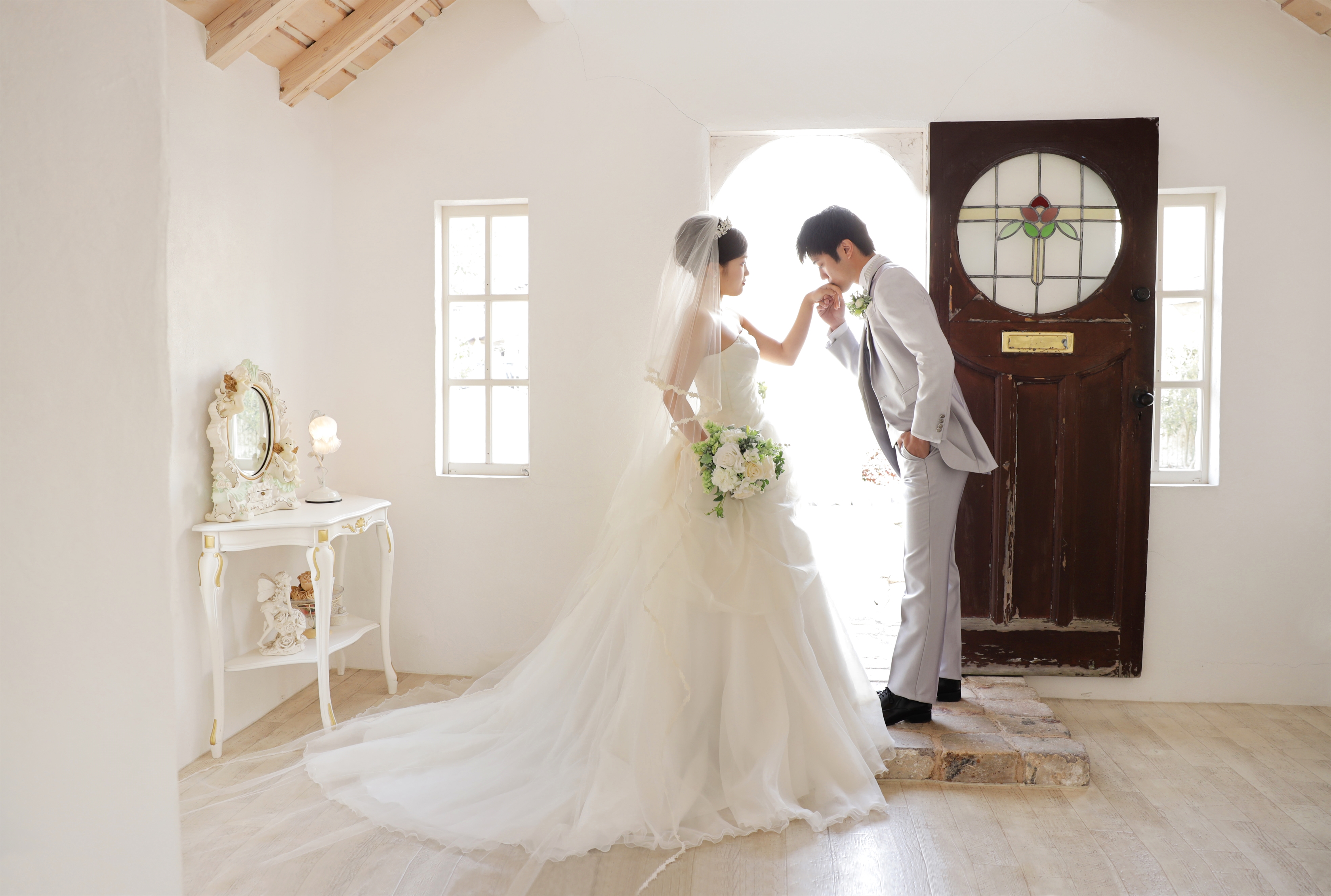 光あふれるチャペル入口で新婦の手にくちづけるタキシード姿の新郎