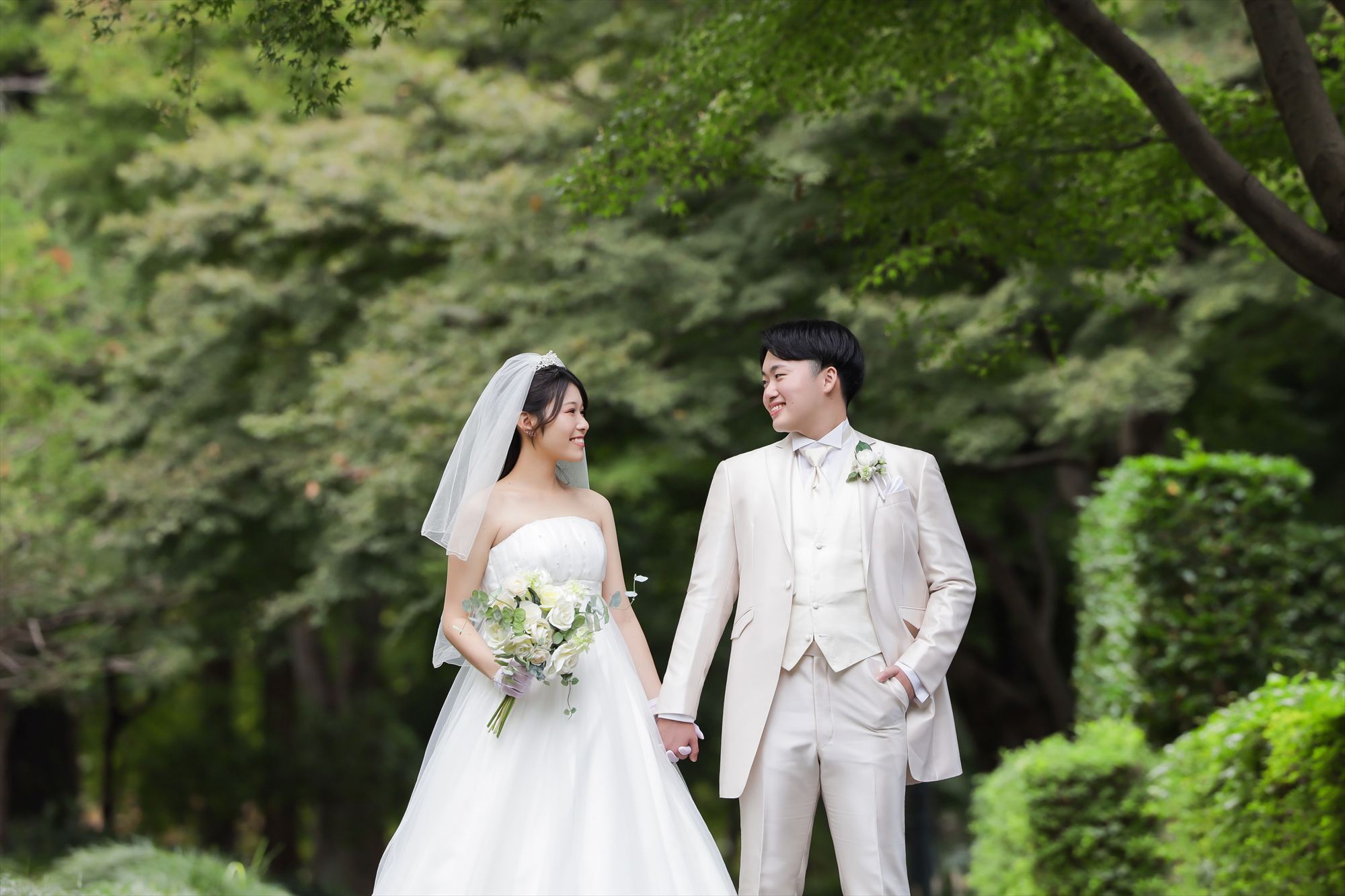 新緑の公園でウェディングドレスと白のタキシード姿で手をつなぐ新郎新婦の洋装ロケーション前撮り