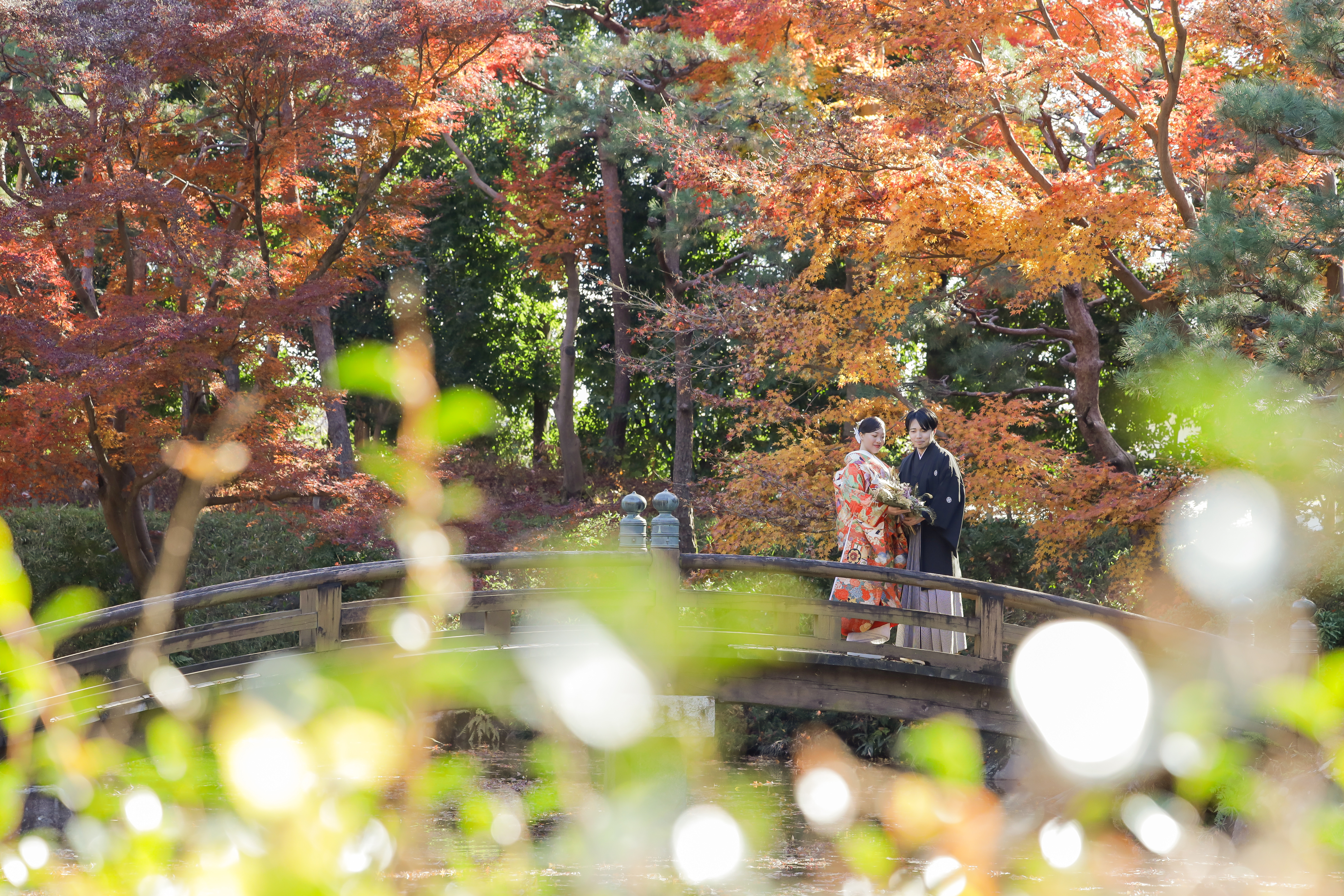 紅葉に包まれた日本庭園の太鼓橋にたたずむ色打掛の新婦と紋付袴の新郎