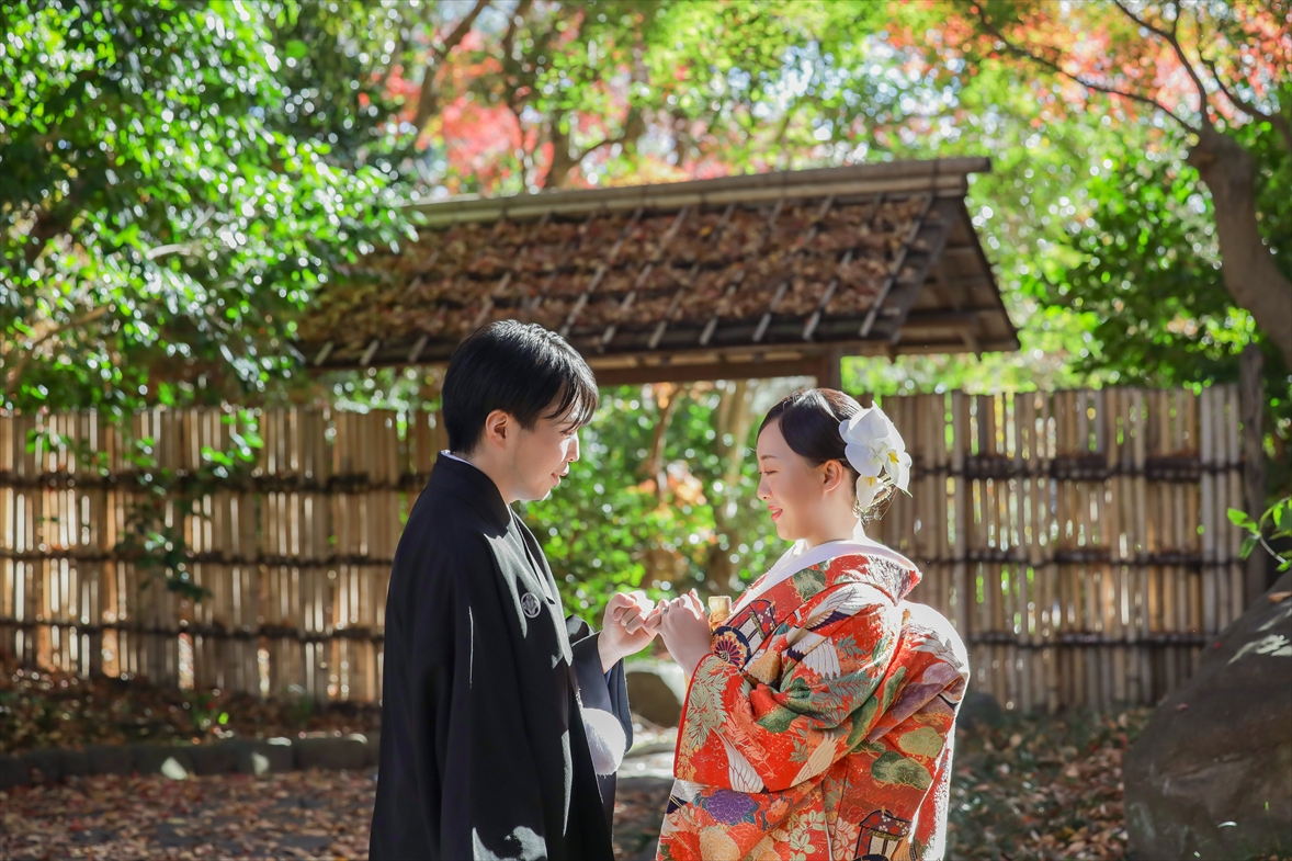 竹垣と茅葺門を背に手を取り合って微笑む朱色の色打掛の新婦と紋付袴の新郎