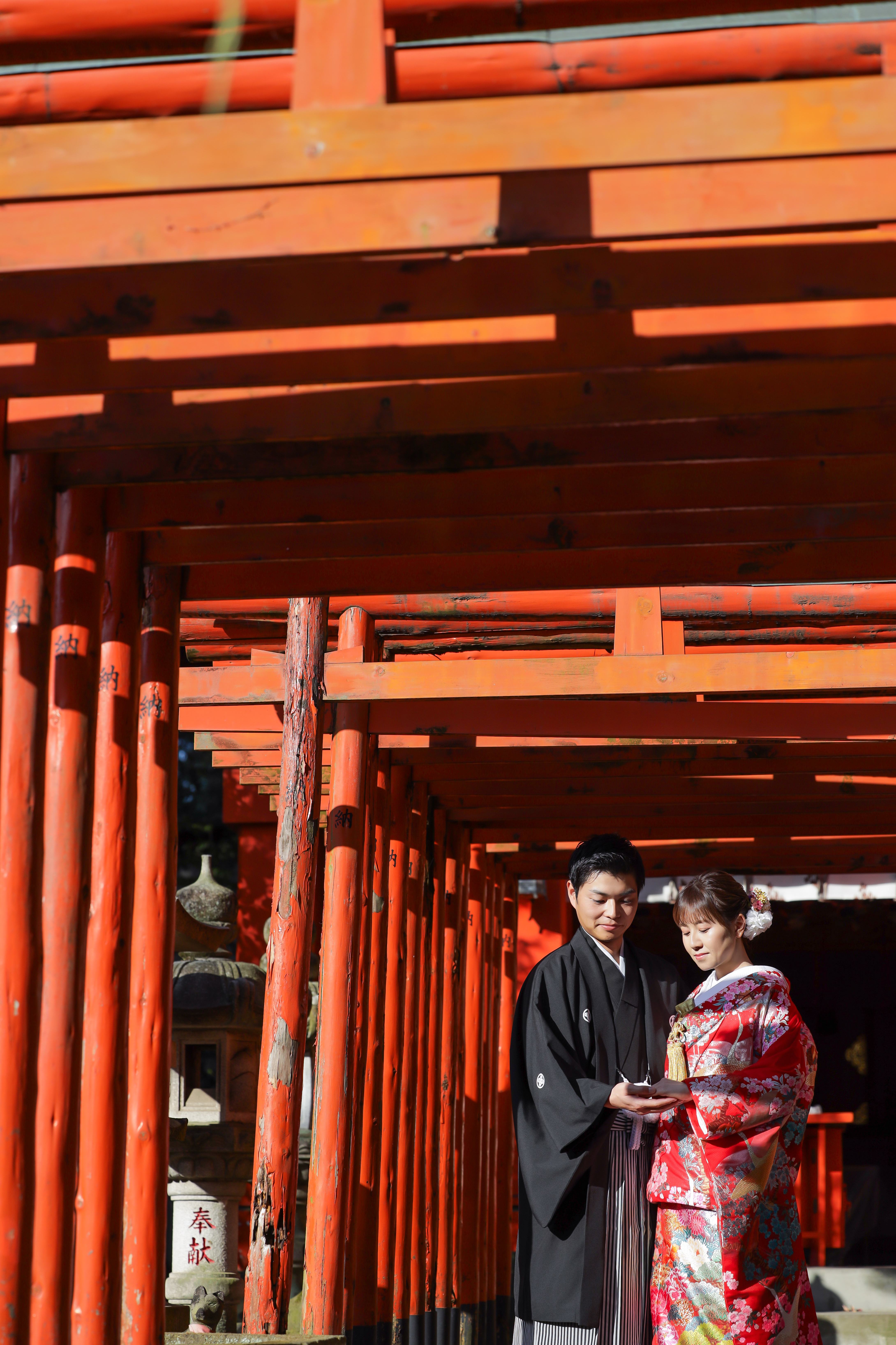 朱色の千本鳥居の下で寄り添う赤い色打掛の新婦と紋付袴の新郎