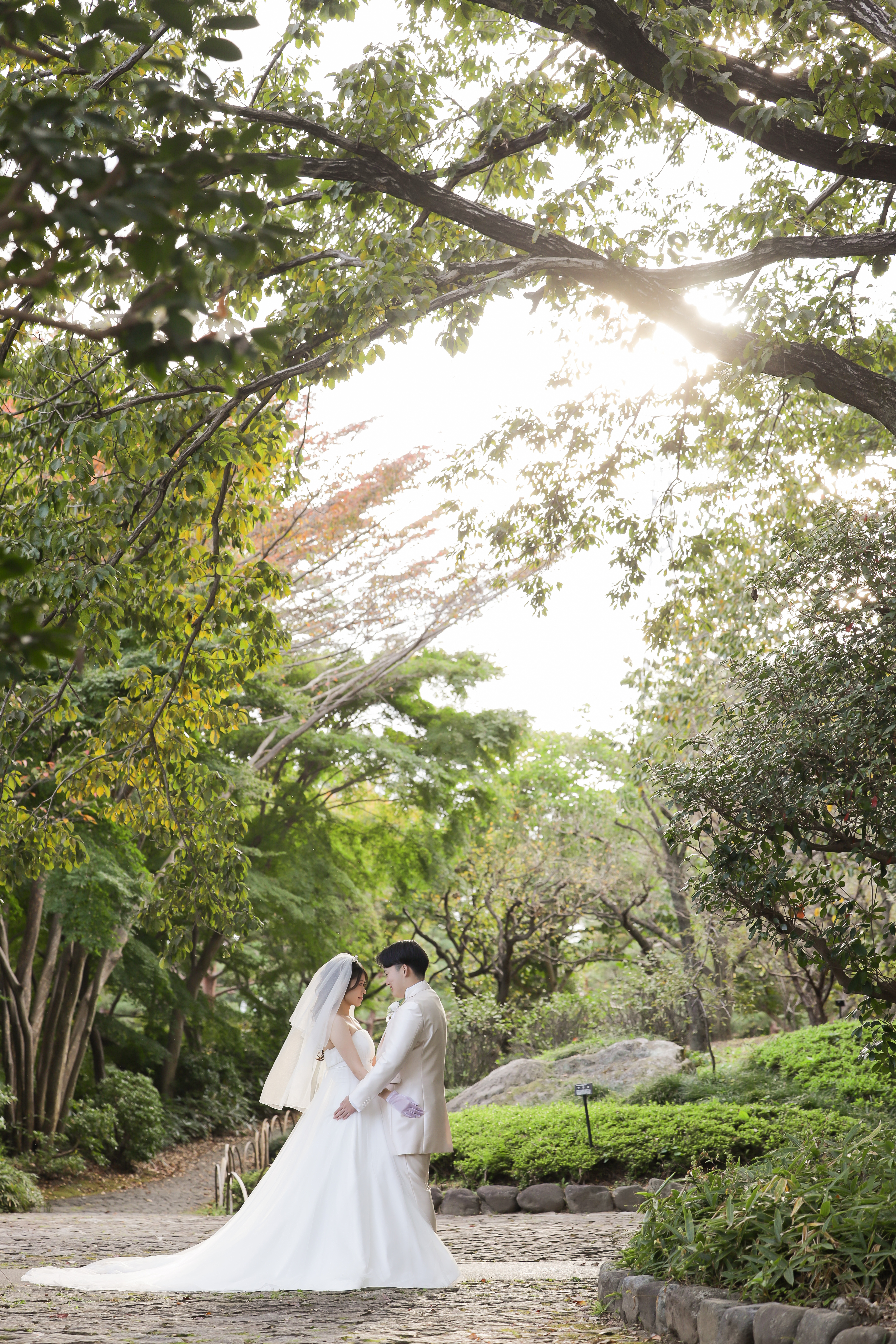 木漏れ日あふれる庭園の石畳で見つめ合うウェディングドレスの新婦と白タキシードの新郎
