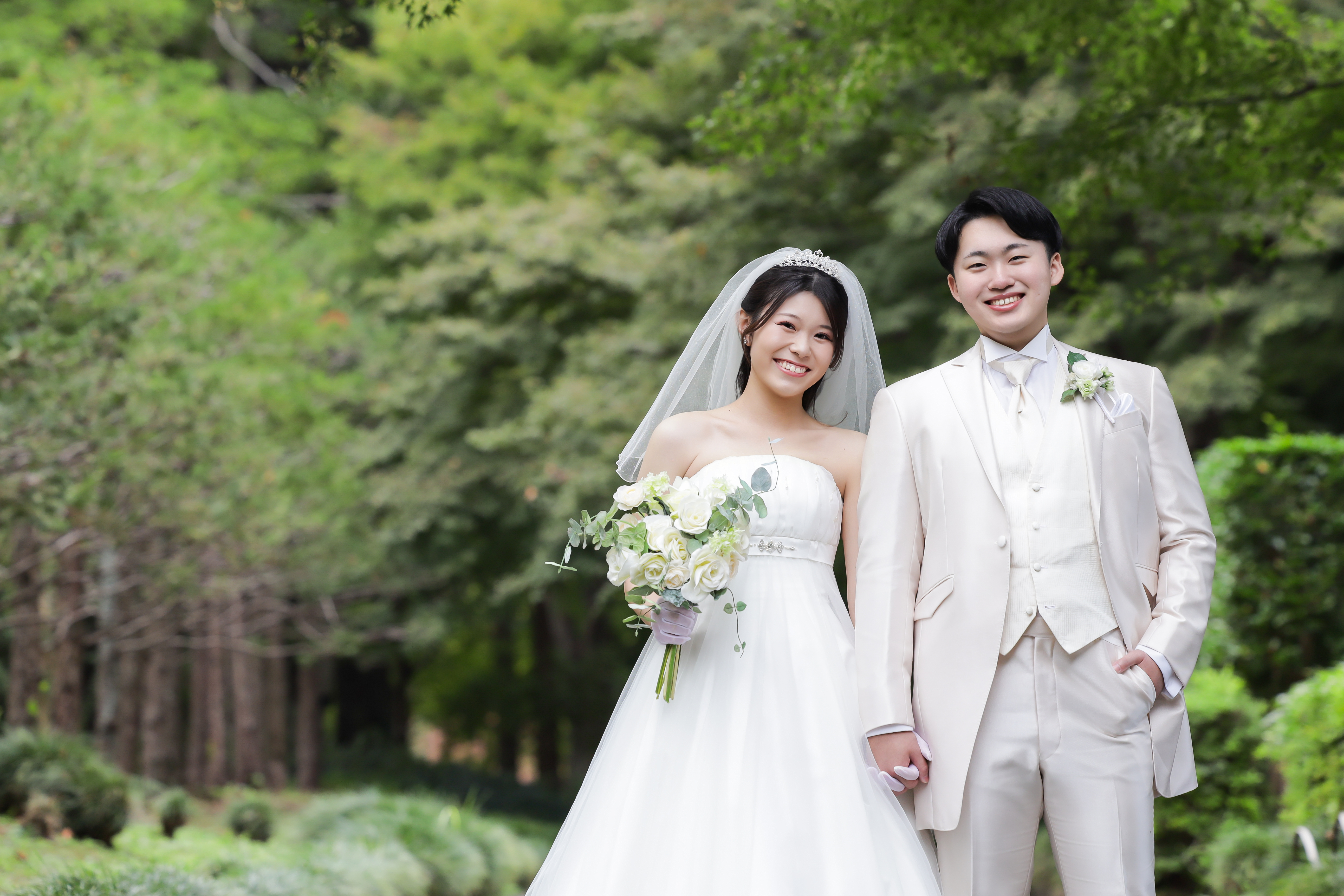 新緑の庭園でブーケを手に手をつなぎ笑顔で並ぶウェディングドレスの新婦と白タキシードの新郎