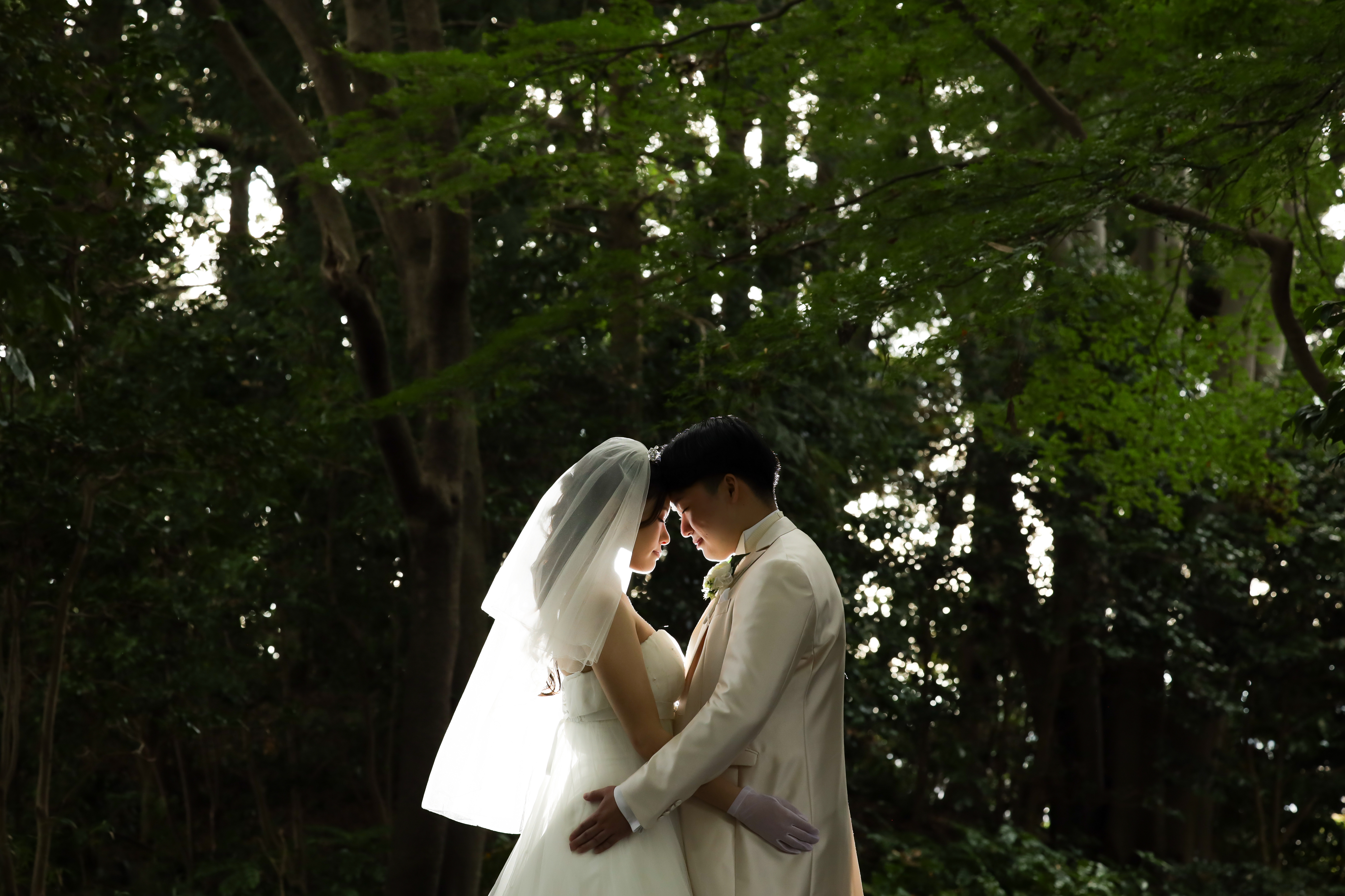 木立の緑を背に額を寄せて寄り添うベール姿の新婦と白タキシードの新郎