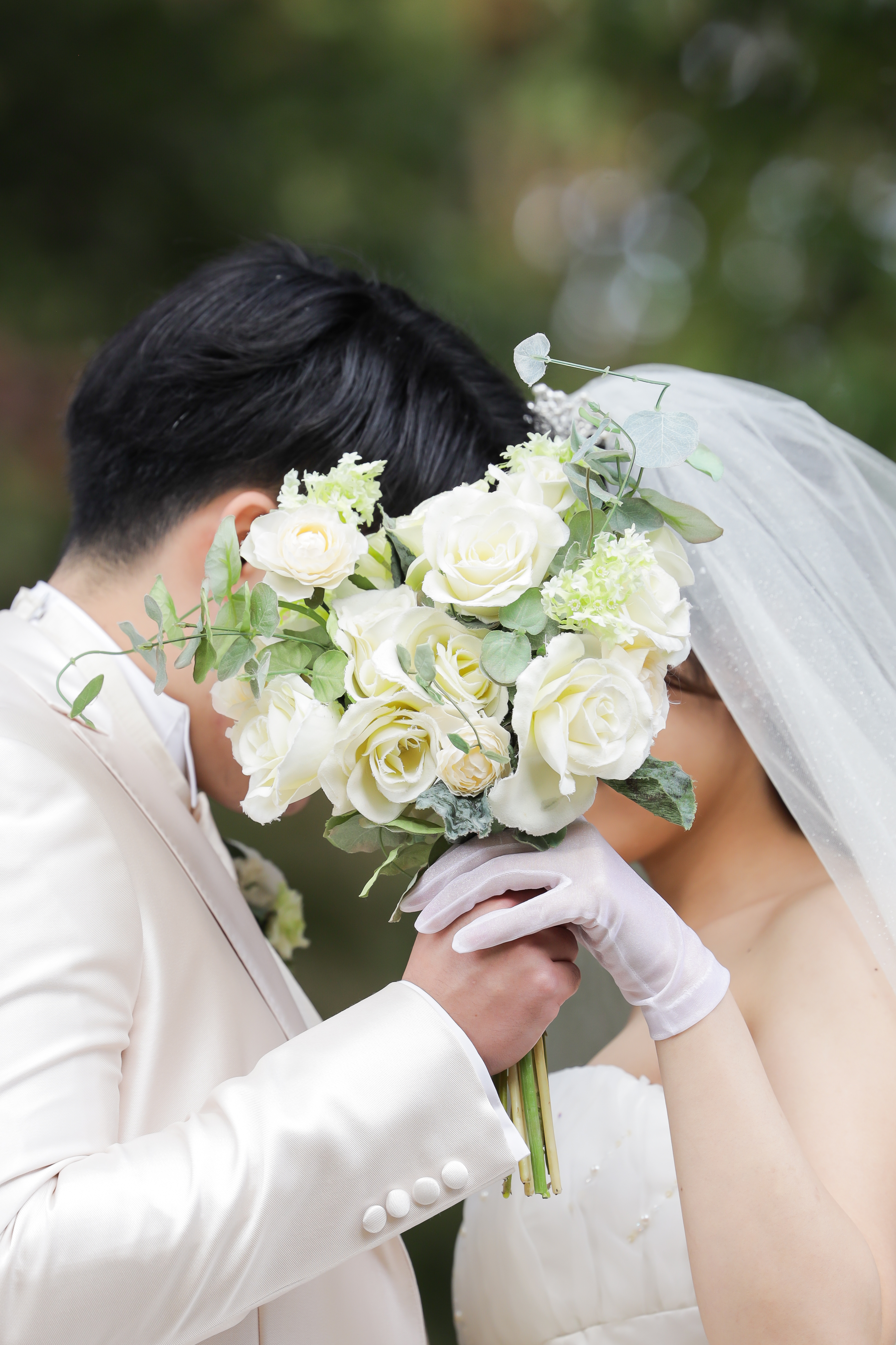 白バラのブーケで顔を隠しキスを交わすベール姿の新婦と白タキシードの新郎