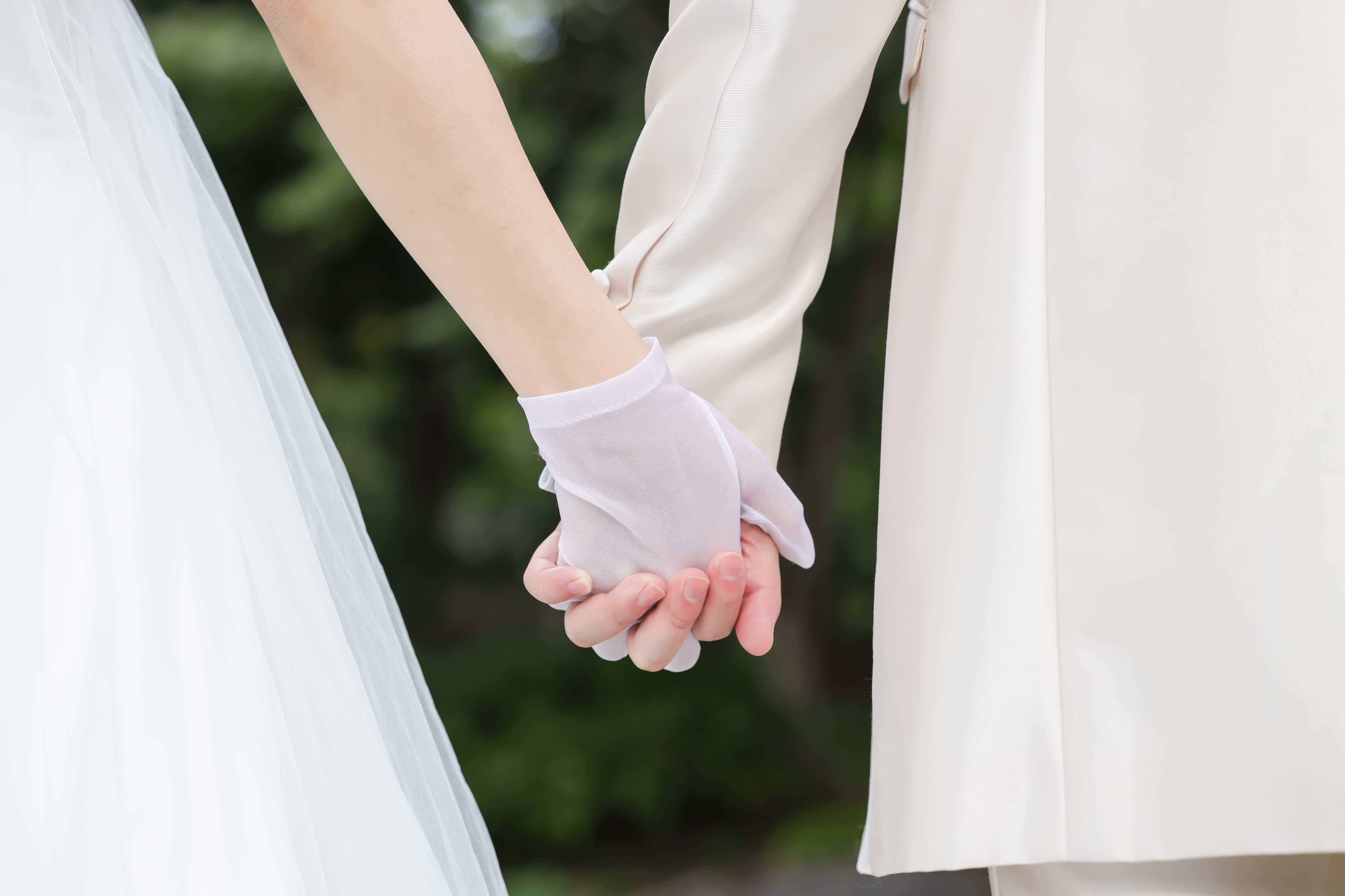 緑を背景に手をつなぐ新郎新婦の手元、白いレースグローブとドレスの裾