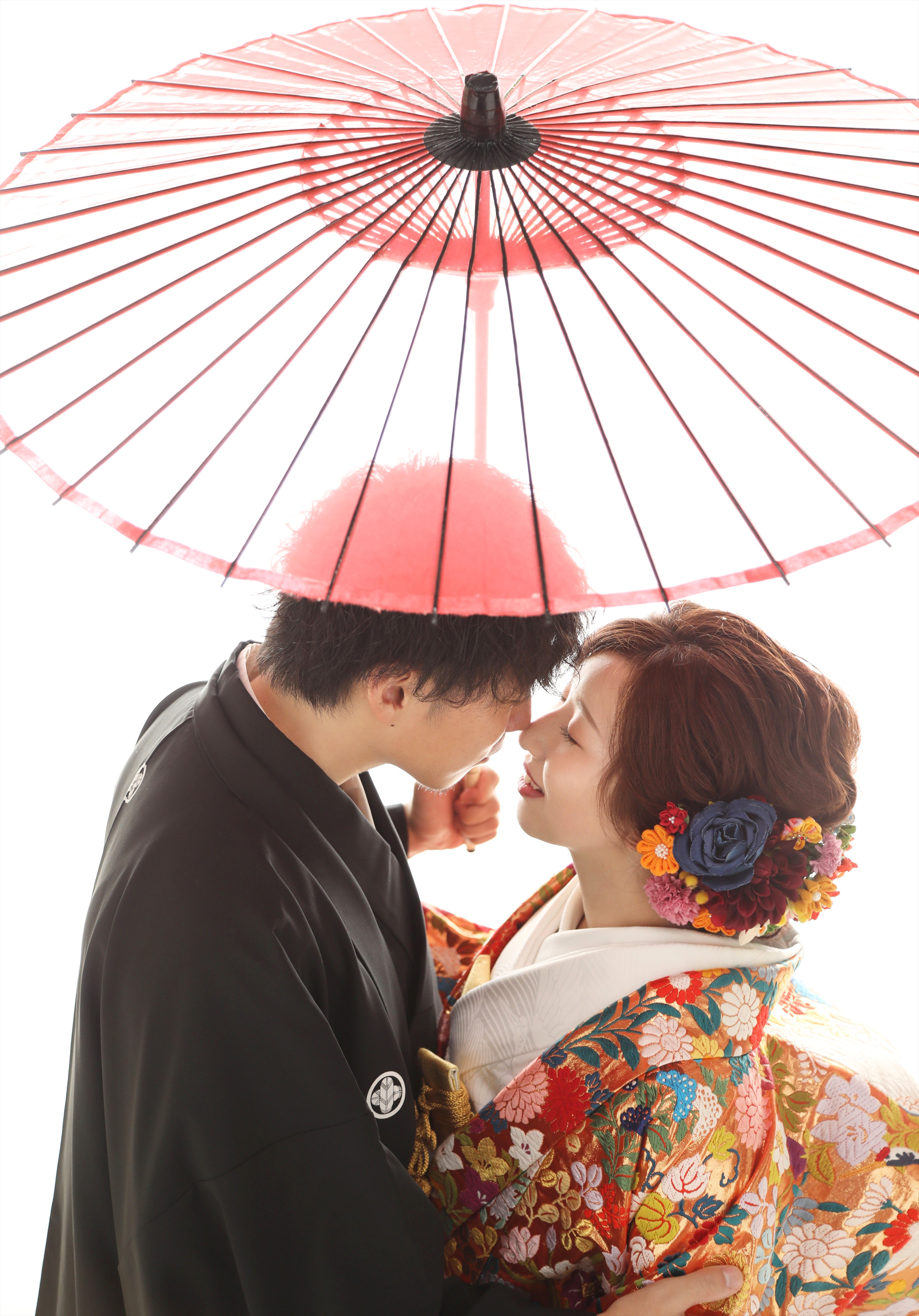 赤い和傘の下で顔を寄せ合う色打掛の新婦と黒紋付袴の新郎