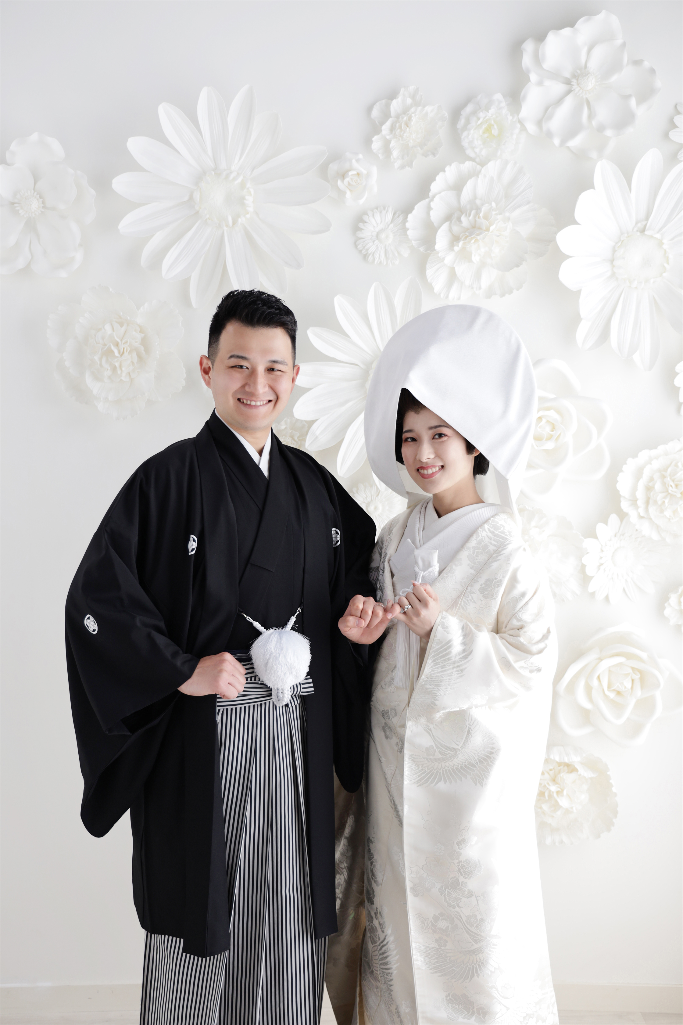白い花の壁を背に腕を組む綿帽子に白無垢の新婦と黒紋付袴の新郎