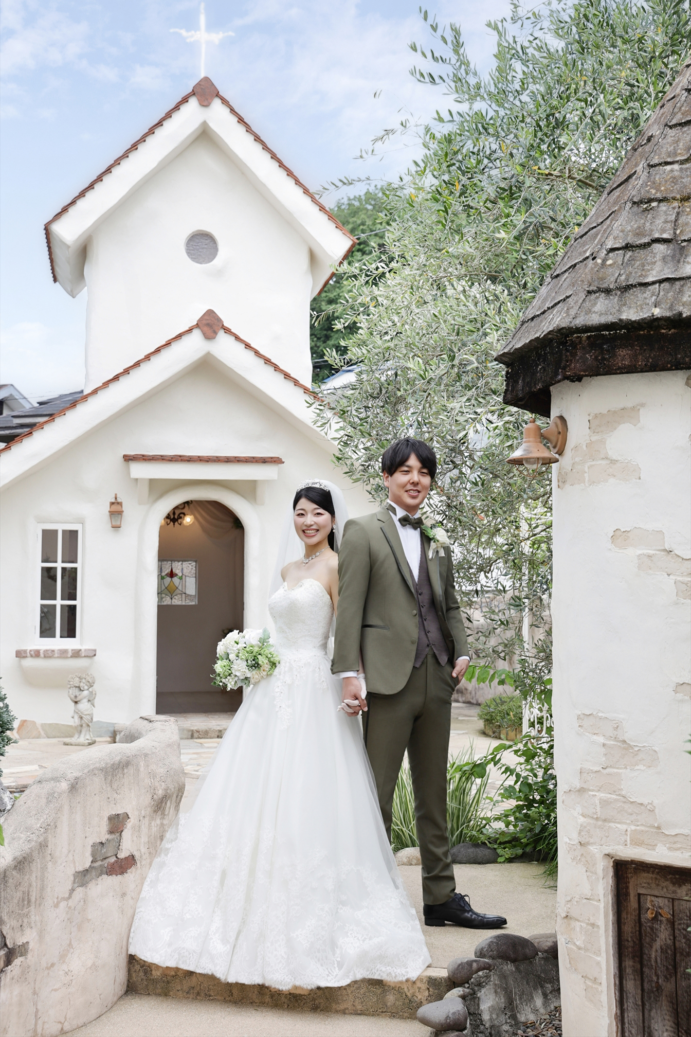 白い教会の前で手をつなぐウェディングドレスの新婦とカーキのタキシードの新郎