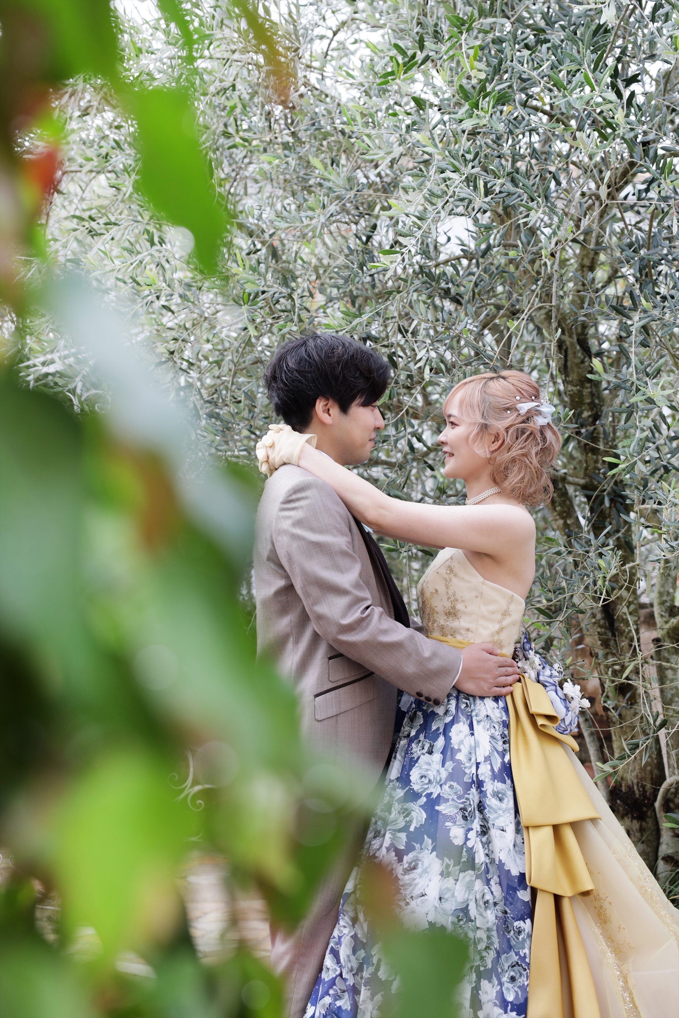 オリーブの木の下で抱き合うブルーの花柄カラードレスの新婦と新郎